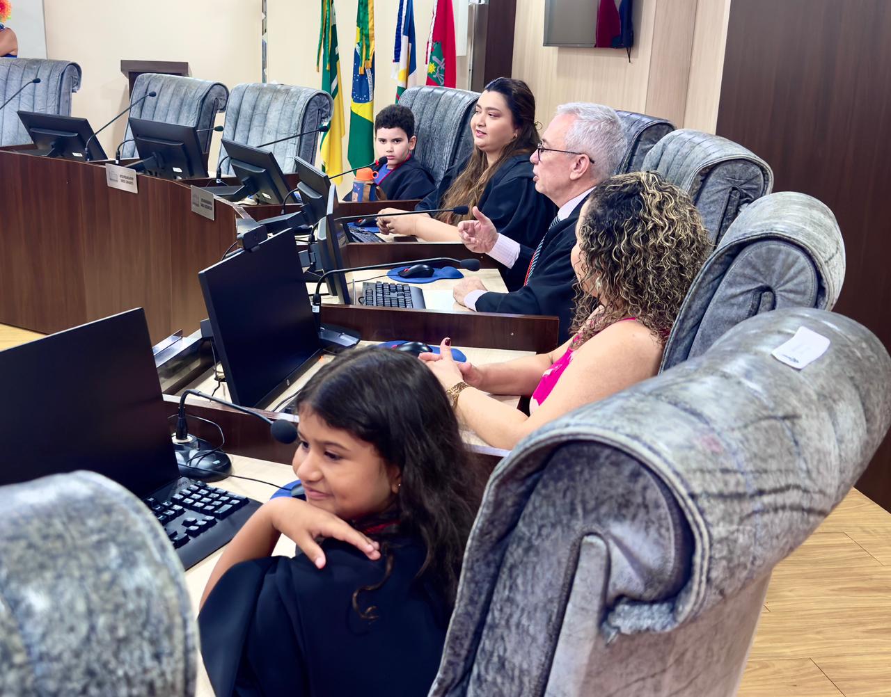  A imagem mostra uma sala de audiências, várias pessoas estão sentadas em cadeiras estofadas de cor cinza, posicionadas em uma mesa semicircular com computadores e microfones. Entre elas, há crianças e adultos, alguns vestidos com capas pretas semelhantes às usadas por juízes. O vice-presidente do TJRR, desembargador Almiro Padilha, fala enquanto gesticula, sendo observado atentamente pelos demais. Ao fundo, estão hasteadas bandeiras do Brasil, de Roraima e do Tribunal de Justiça.