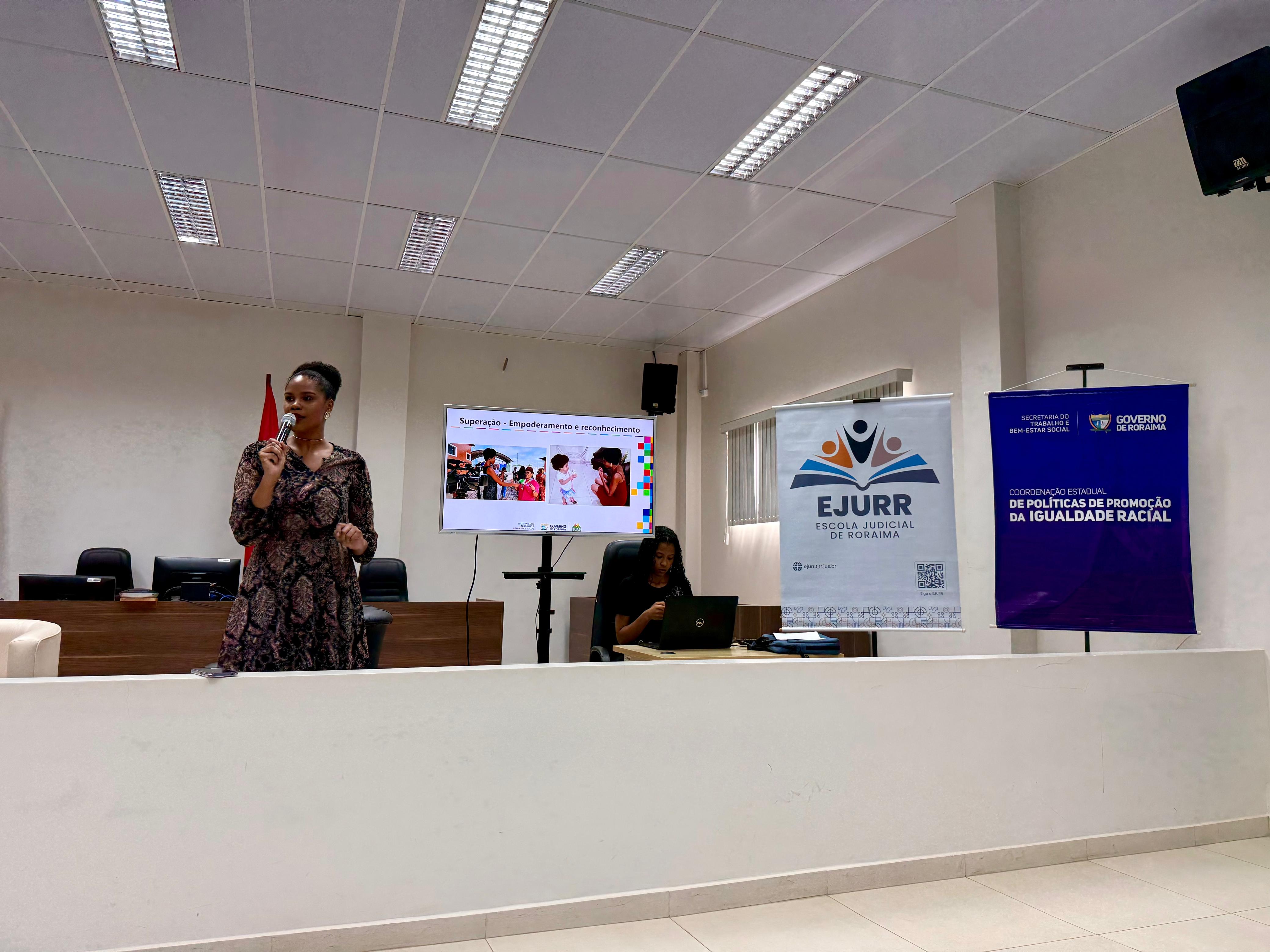 A coordenadora de Políticas de Promoção da Igualdade Racial da Setrabes, Rafaela André, durante apresentação em um salão com banners sobre igualdade racial. Outra mulher está sentada ao fundo, usando um laptop.