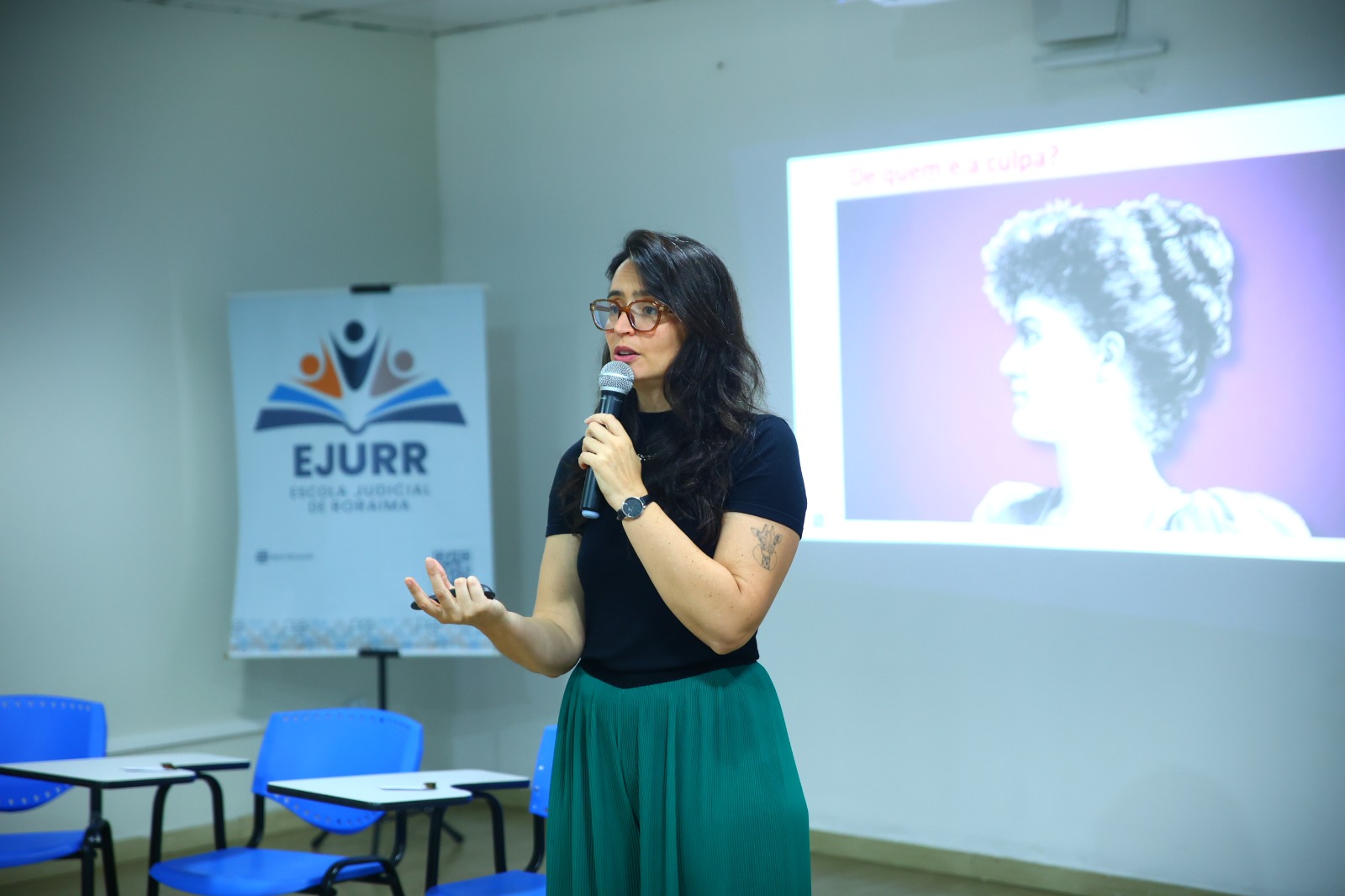 imagem dando close na palestrante em um momento de fala, segurando um microfone ao fundo há o banner da ejurr. imagem dando close na palestrante em um momento de fala, segurando um microfone ao fundo há o banner da ejurr.