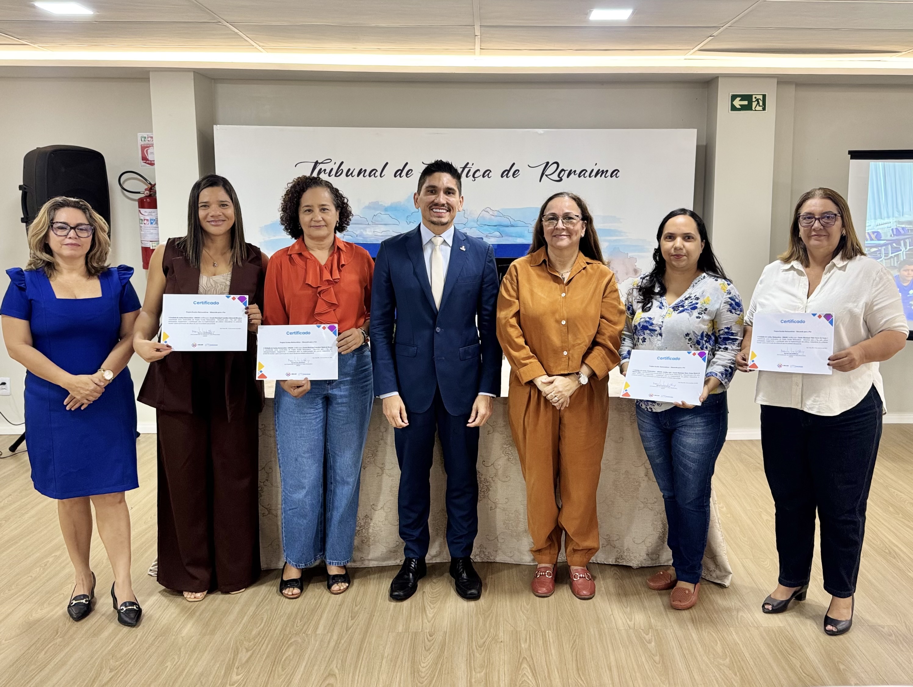 Imagem colorida mostra sete pessoas em pé, lado a lado, em um ambiente interno. Três delas seguram certificados nas mãos. Ao fundo, há um painel com a inscrição "Tribunal de Justiça de Roraima" e paisagem ilustrada. Todas estão vestidas formalmente, com roupas sociais.