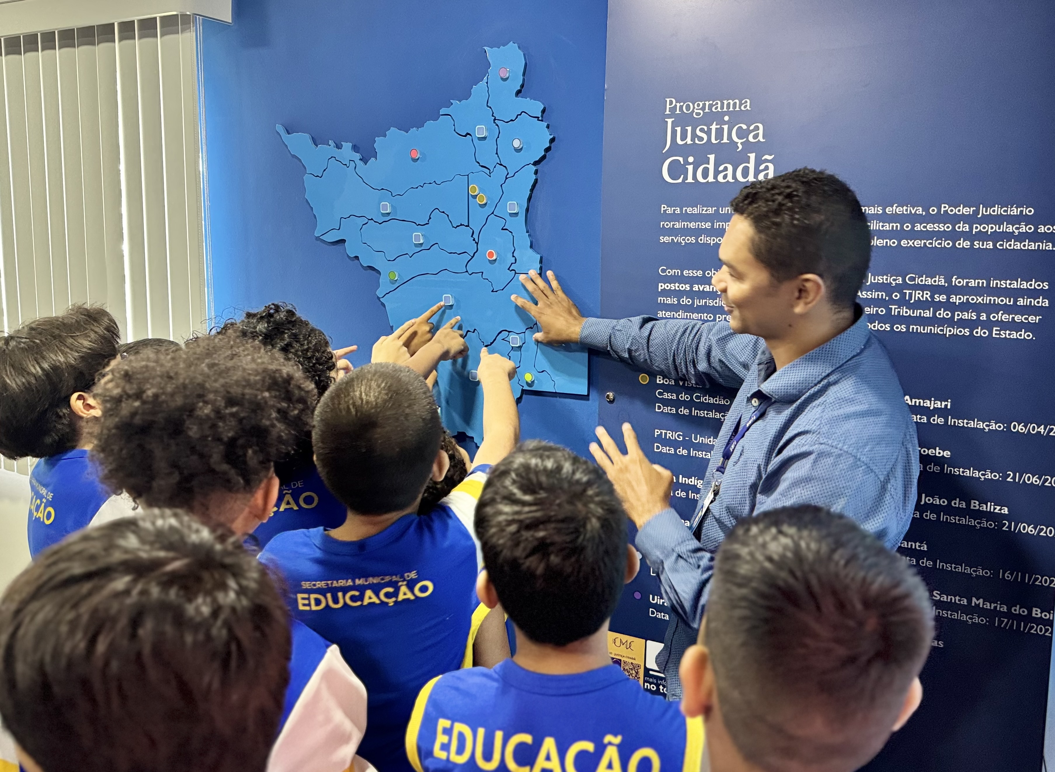 Imagem colorida mostra um grupo de crianças de camiseta azul e amarelo, acompanhadas por um homem adulto. Elas estão reunidas em frente a um painel azul com a ilustração de um mapa do estado de Roraima, apontando para diferentes regiões.