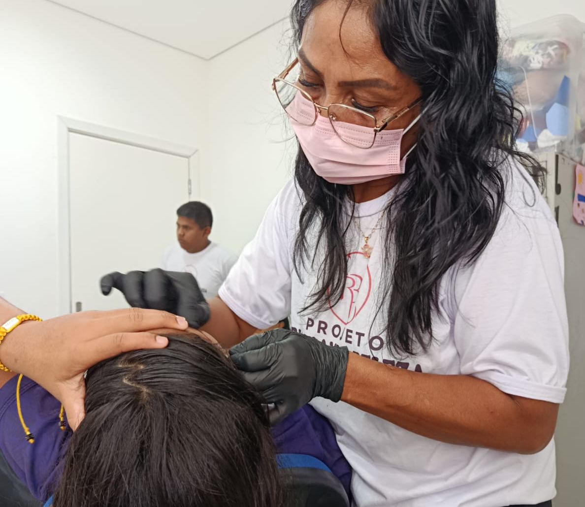 Imagem colorida mostra uma mulher usando luvas pretas manipulando o cabelo de outra pessoa que está sentada à sua frente. A mulher veste uma camiseta branca estampada com o texto "PROJETO" e parte de um logo visível. Ao fundo, aparece um homem desfocado, também de camiseta branca, e detalhes do ambiente, como uma porta branca e prateleiras.