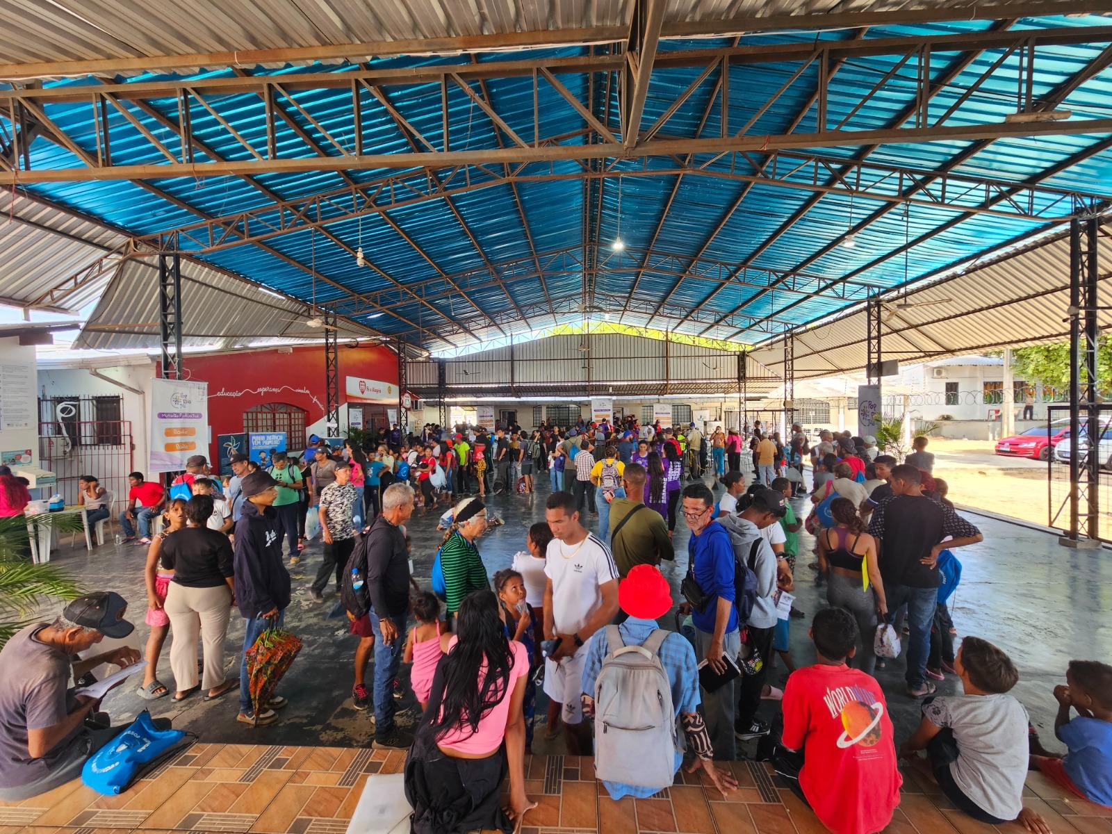 Multidão de pessoas em um espaço coberto, aguardando em filas. O ambiente apresenta um teto azul e paredes com informações visíveis ao fundo.