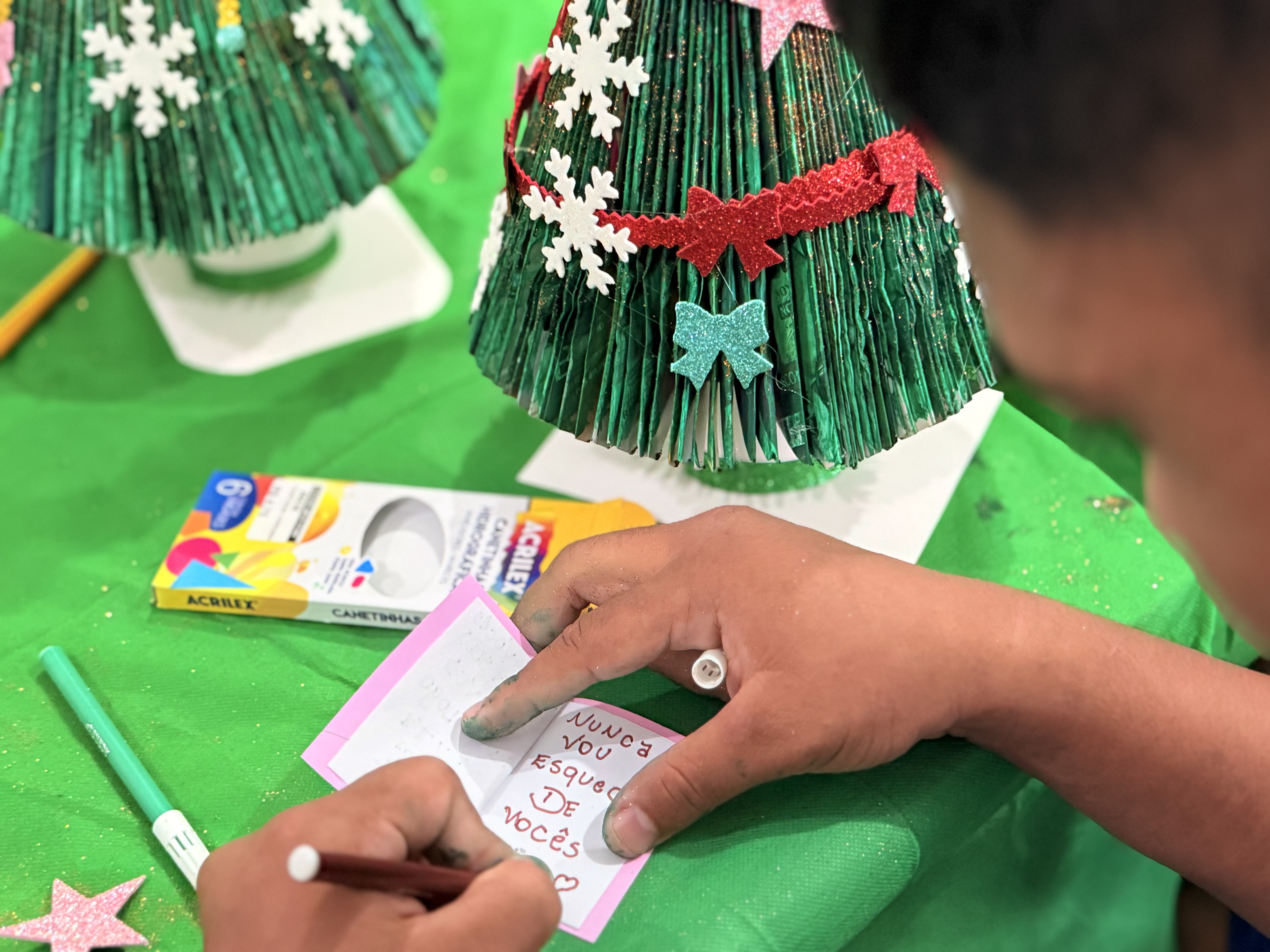 Imagem colorida mostra uma pessoa escrevendo em um cartão de papel, próximo a uma árvore de Natal artesanal decorada. Sobre a mesa verde, há uma caixa de canetinhas coloridas e outros cartões, e é possível ler a frase "Nunca vou esquecer de vocês" escrita no cartão.