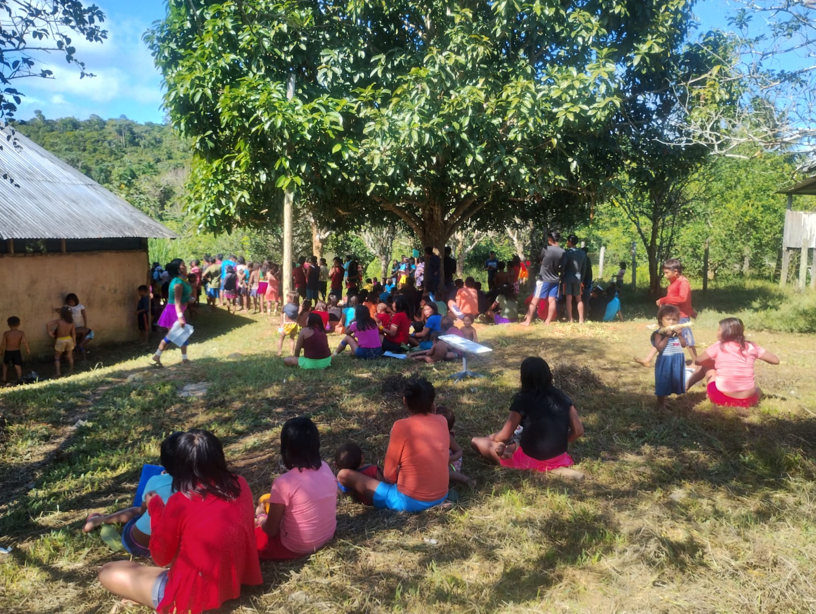 Imagem colorida mostra uma grande área ao ar livre, com gramado e árvores, onde várias pessoas, principalmente crianças, estão sentadas ou em pé. Algumas brincam ou conversam próximas a uma casa com telhado de zinco. O dia está ensolarado, e o ambiente transmite sensação de comunidade e encontro coletivo. Muitas pessoas vestem roupas coloridas e os rostos estão desfocados.