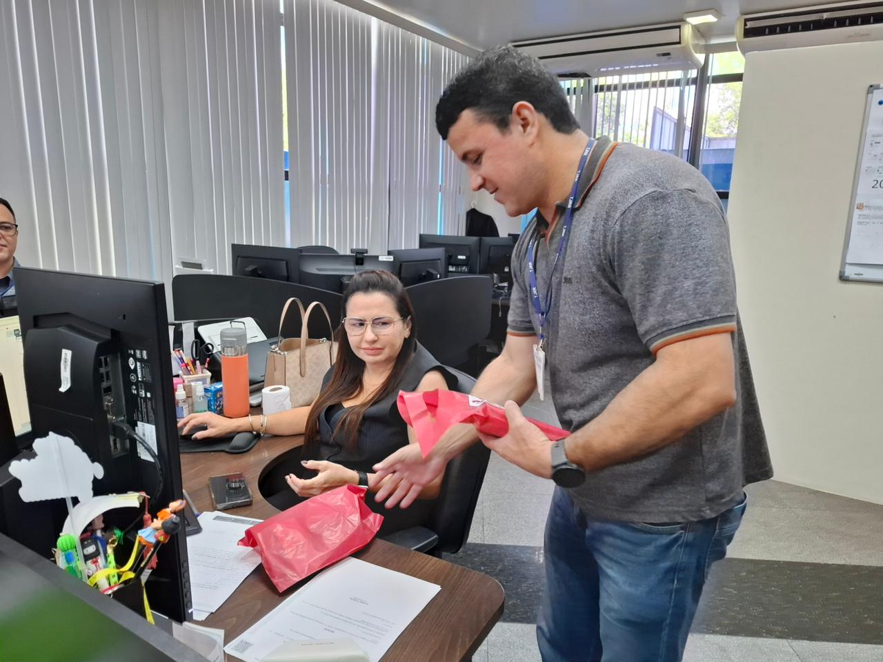 Imagem colorida mostra um ambiente com computadores e objetos pessoais sobre as mesas. Um homem em pé, usando crachá, entrega um pacote vermelho para uma mulher sentada à frente do computador. Ao fundo, outro homem aparece enviado, também em frente a um computador.