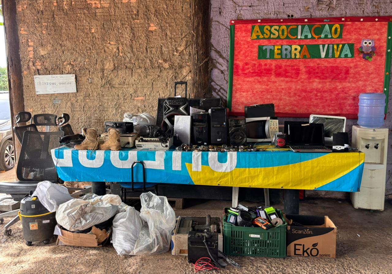 Mesa coberta por toalha azul e amarela com vários bens eletrônicos e objetos. Há computadores, aparelhos de som, ferramentas, cabos e outros itens. Ao fundo, vê-se o cartaz “Associação Terra Viva” e o ambiente simples do galpão.