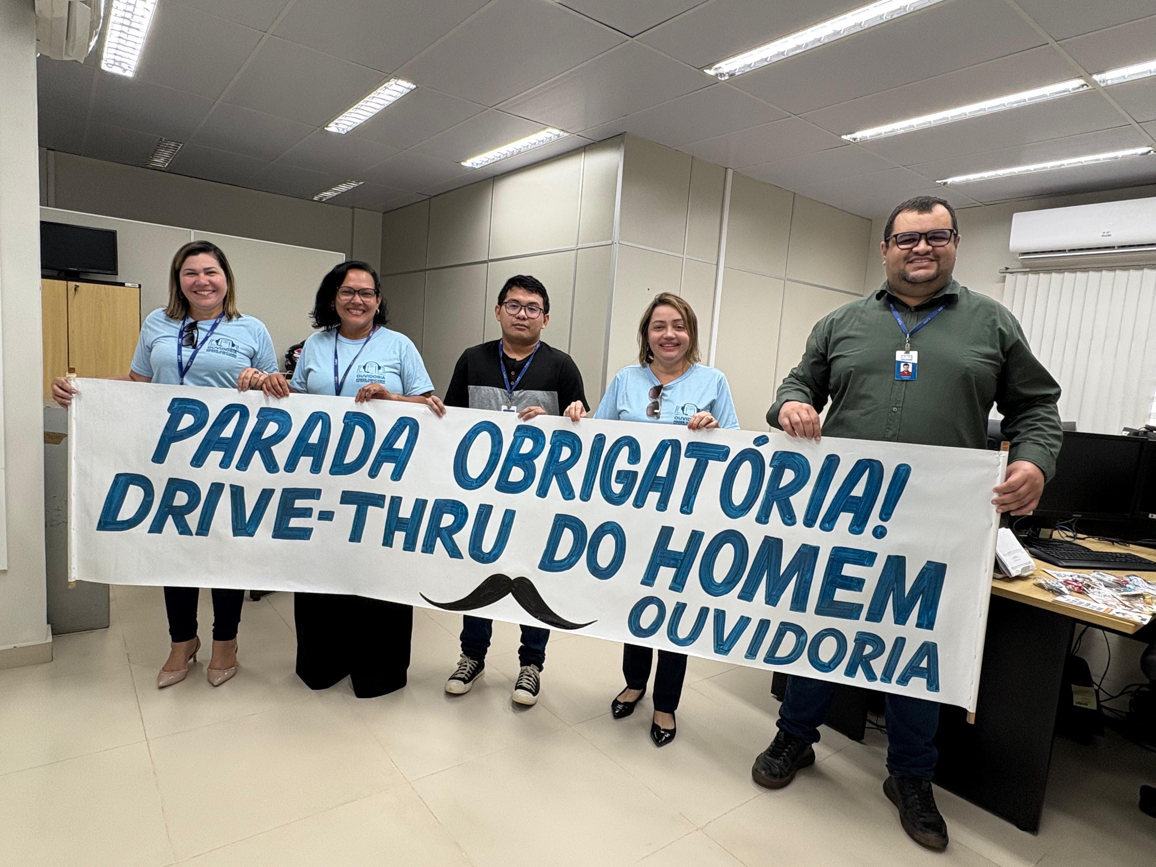 imagem colorida mostra 5 pessoas, sendo 2 mulheres e 3 homens segurando uma faixa com o texto "Parada obrigatória Drive thru do homem ouvidoria". imagem colorida mostra 5 pessoas, sendo 2 mulheres e 3 homens segurando uma faixa com o texto "Parada obrigatória Drive thru do homem ouvidoria".