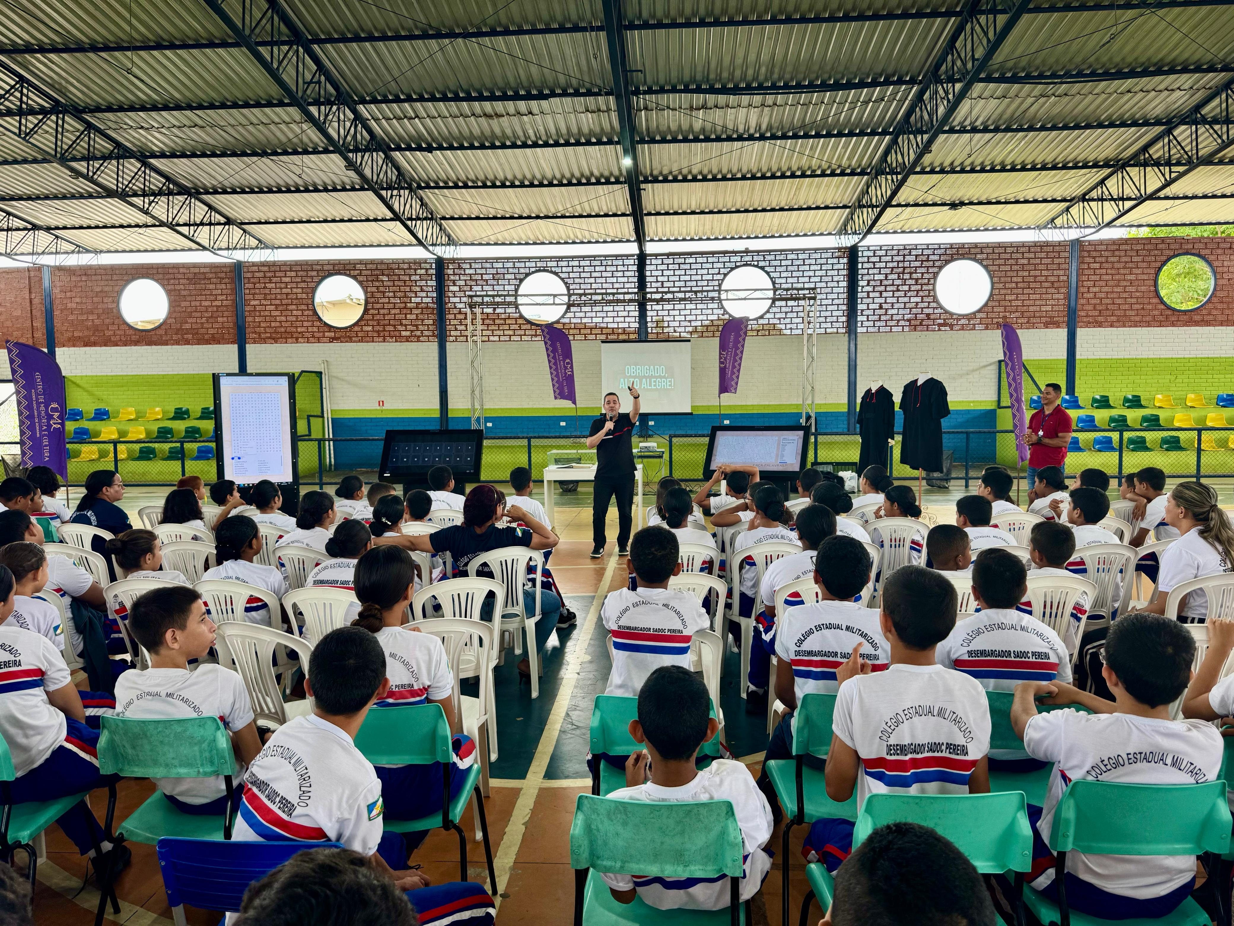 A imagem colorida mostra um grupo de jovens, a maioria vestindo camisetas brancas com logotipos coloridos, sentados em cadeiras brancas e verdes, de costas para a câmera. Eles estão em um espaço amplo, que parece ser um ginásio ou auditório, com teto alto e vigas metálicas. À frente da plateia, um homem em pé, no centro, gesticula enquanto se dirige ao público. Ao seu lado, há telões e, à direita, algumas pessoas em pé e cadeiras vazias.