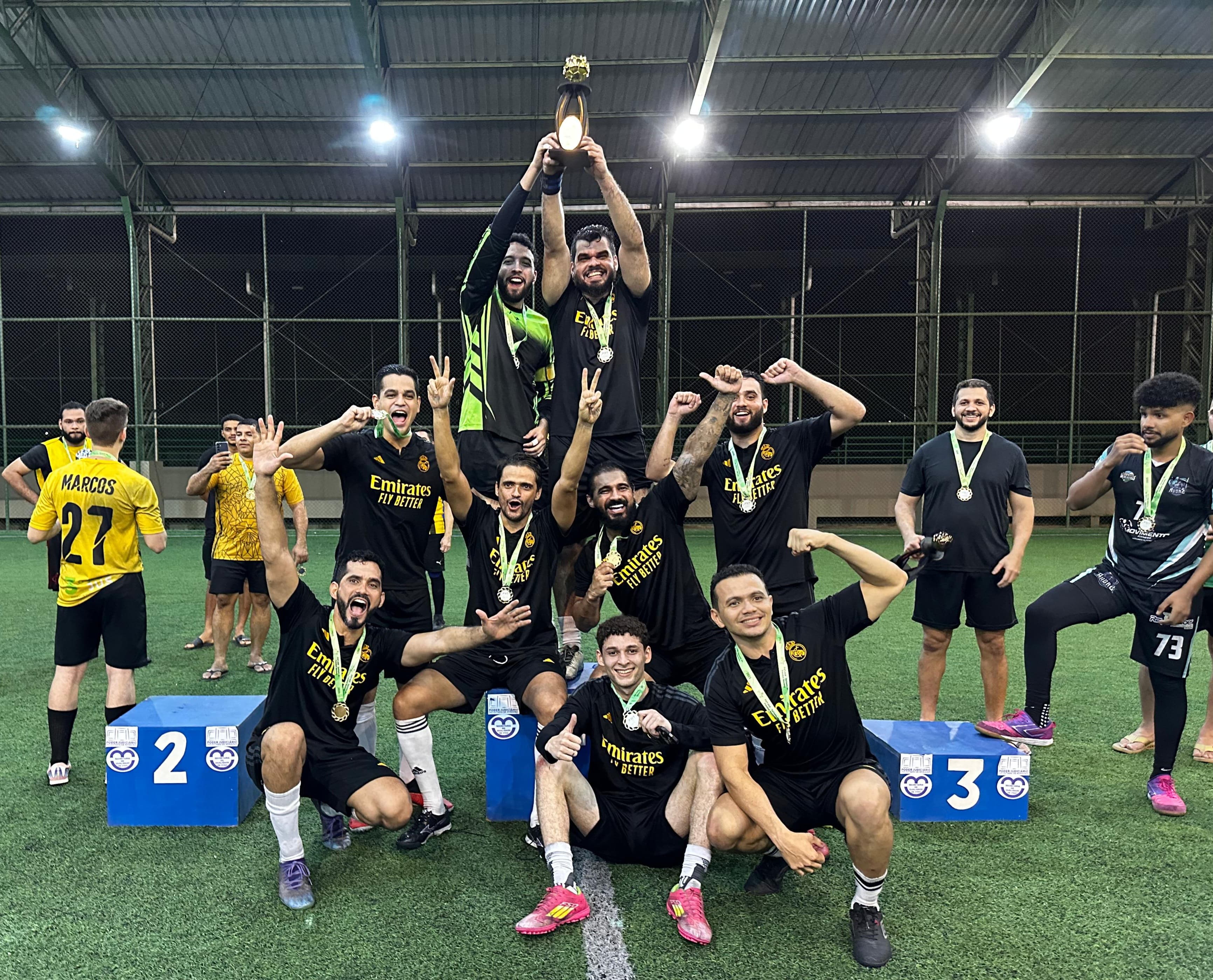 Imagem colorida mostra o time preto celebrando a vitória em um campo de grama sintética. No centro da imagem, dois jogadores, posicionados em cima de um pódio, erguem uma grande taça dourada, enquanto os demais companheiros de equipe, exibindo medalhas, os rodeiam em poses de comemoração. Há pódios numerados ("2" e "3") visíveis na parte inferior da foto. O ambiente é uma quadra coberta, com iluminação artificial. Imagem colorida mostra o time preto celebrando a vitória em um campo de grama sintética. No centro da imagem, dois jogadores, posicionados em cima de um pódio, erguem uma grande taça dourada, enquanto os demais companheiros de equipe, exibindo medalhas, os rodeiam em poses de comemoração. Há pódios numerados ("2" e "3") visíveis na parte inferior da foto. O ambiente é uma quadra coberta, com iluminação artificial.