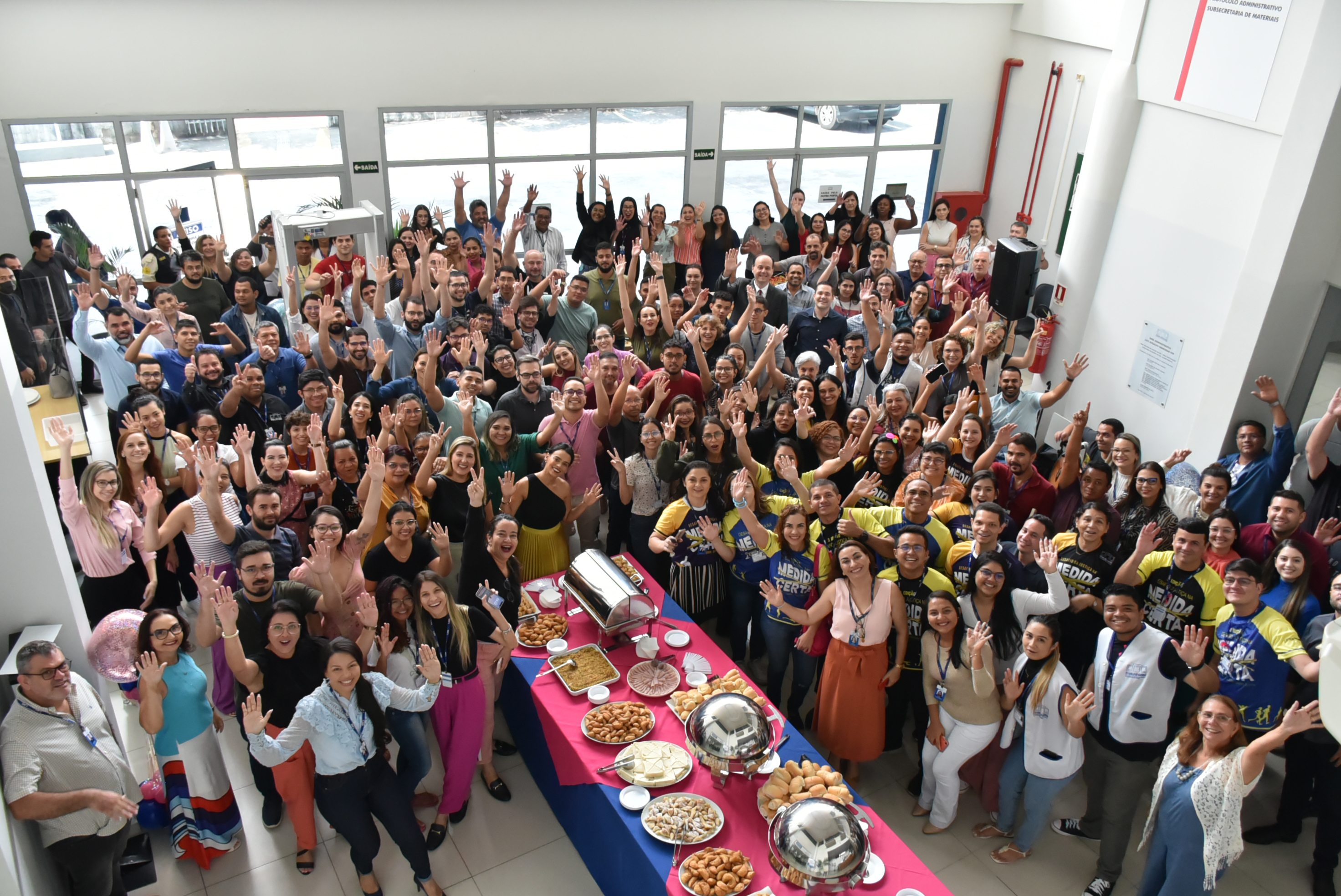 Imagem aérea colorida mostra a sala de recepção da sede administrativa com magistrados, magistradas, servidores, servidoras, terceirizados, terceirizadas,  estagiários e  estagiárias posando para a foto com os braços erguidos. No centro da foto há uma mesa posta com comidas diversificadas.