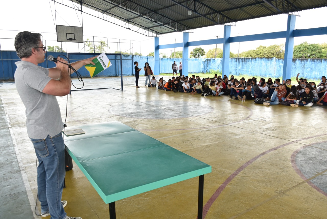 Conselheiro do CNJ, com uma Constituição Federal na mão, discursa para alunos, que estão sentados no chão do pátio da escola