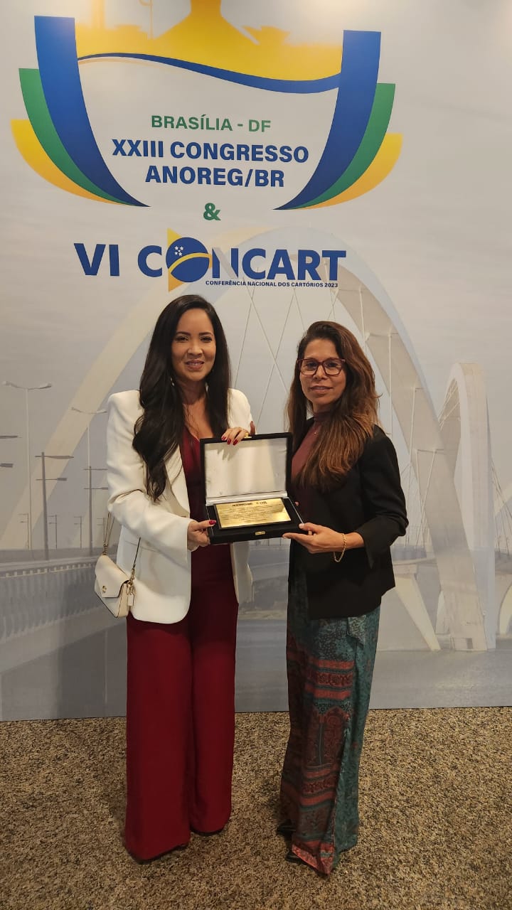 Foto colorida mostra a servidora do TJRR, Luciana Menezes, recebendo premiação em homenagem ao desembargador Mozarildo Cavalcanti, durante congresso nacional Anoreg/BR e posando para foto ao lado da presidente da Associação dos Notários e Registradores do Estado de Roraima (ANOREG/RR), Inês Maraschin.