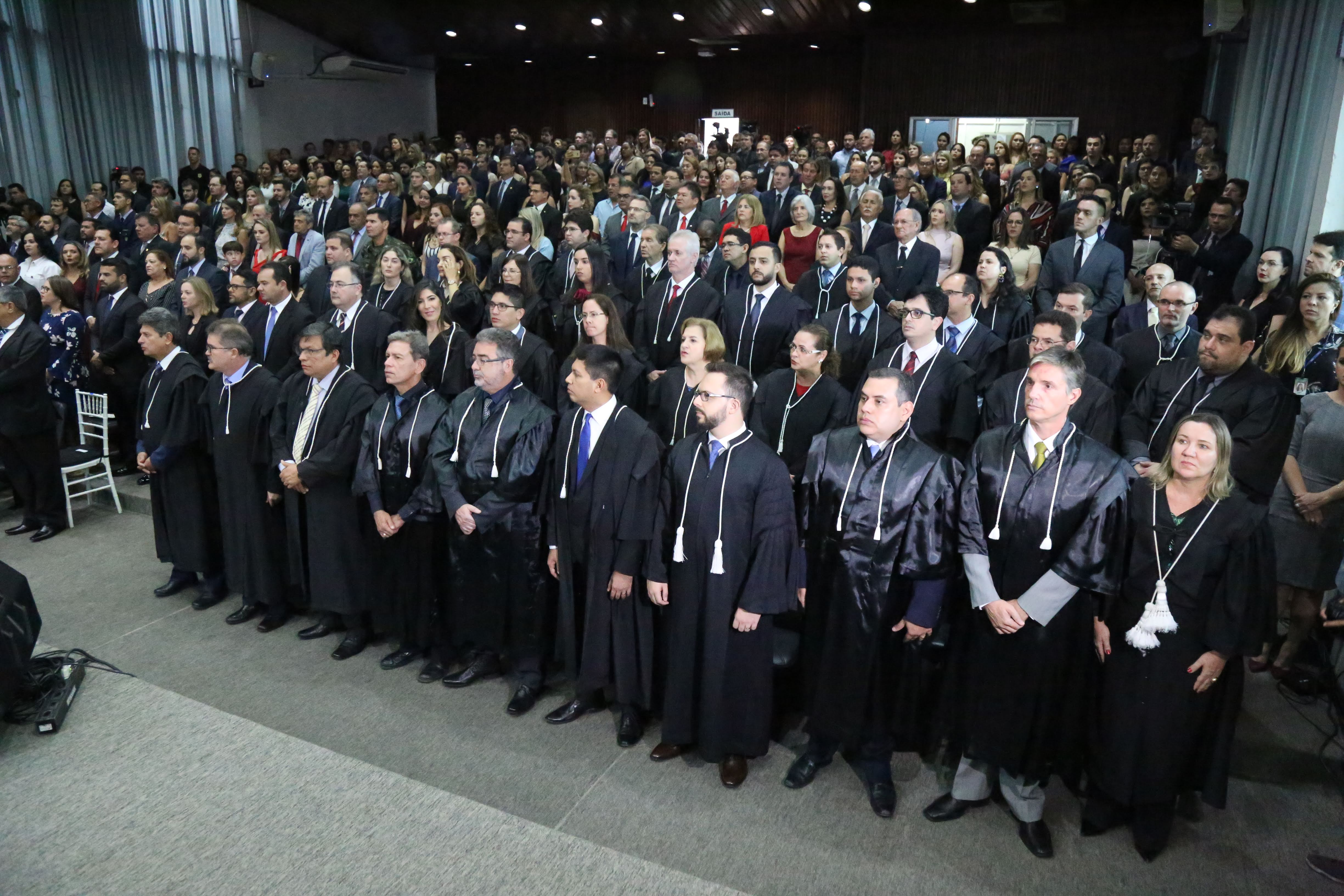 Foto colorida mostra pessoas em pé olhando para o lado esquerdo em um auditório.