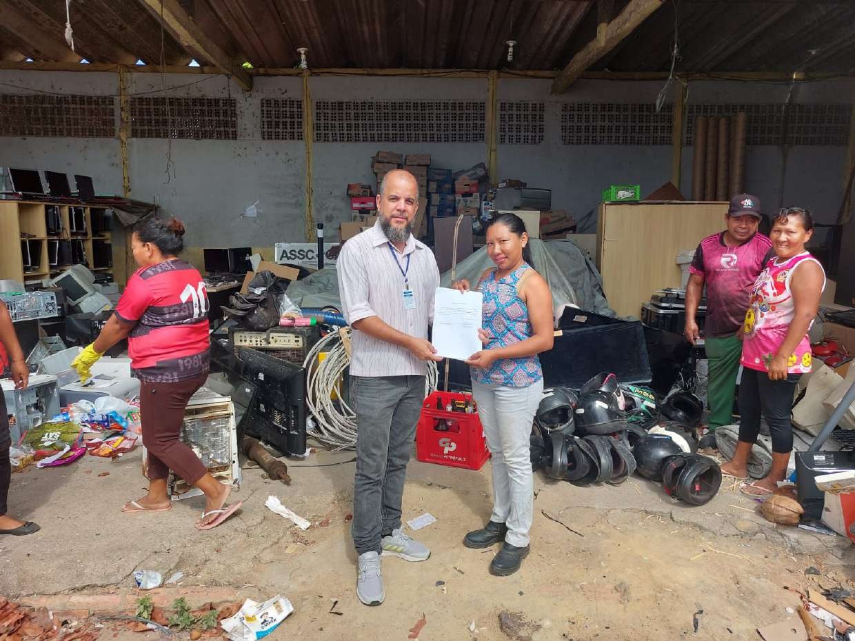 Foto colorida mostra cinco pessoas, na associação Catadores de Materiais Recicláveis Terra Viva, sendo duas delas posando para fotografia com documento assinado em mãos.  Ao redor vários materiais reciclados.