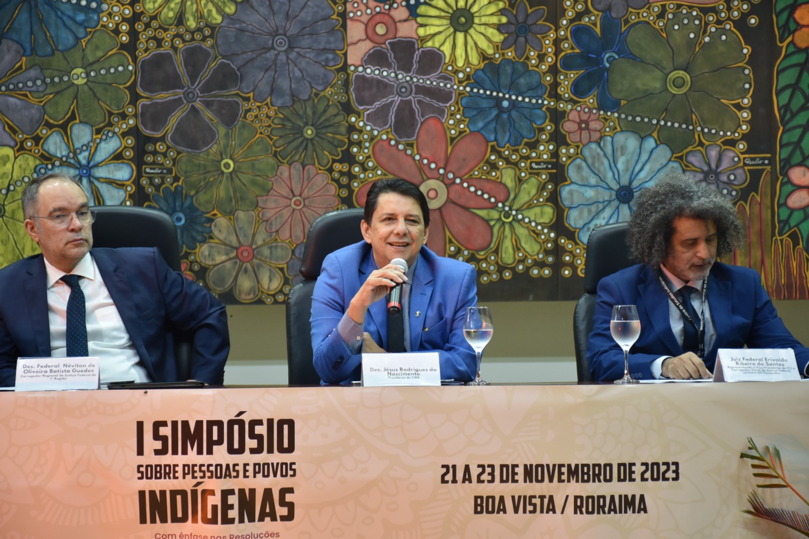 Foto colorida mostra o presidente do Poder Judiciário de Roraima, desembargador Jésus Nascimento, sentado com o microfone na mão, ao lado do Corregedor Regional de Justiça Federal da 1ª Região, desembargador, Néviton Guedes e do o juiz federal Erivaldo Santos, o secretário nacional de acesso à justiça, Marivaldo Pereira. No fundo há um quadro com diversas flores coloridas.