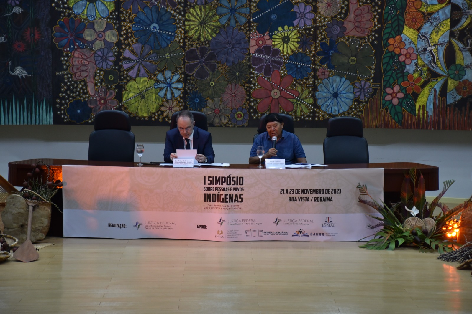 Foto colorida mostra o xamã e líder político do povo Yanomami, Davi Kopenawa, sentado com o microfone na mão, ao lado do Corregedor Regional de Justiça Federal da 1ª Região, desembargador, Néviton Guedes, que está com um papel. No fundo há um quadro com diversas flores coloridas. A frente há uma faixa escrito “ I Simpósio Sobre Pessoas e Povos Indígenas”, “21 a 23 de novembro de 2023, Boa Vista / Roraima”.