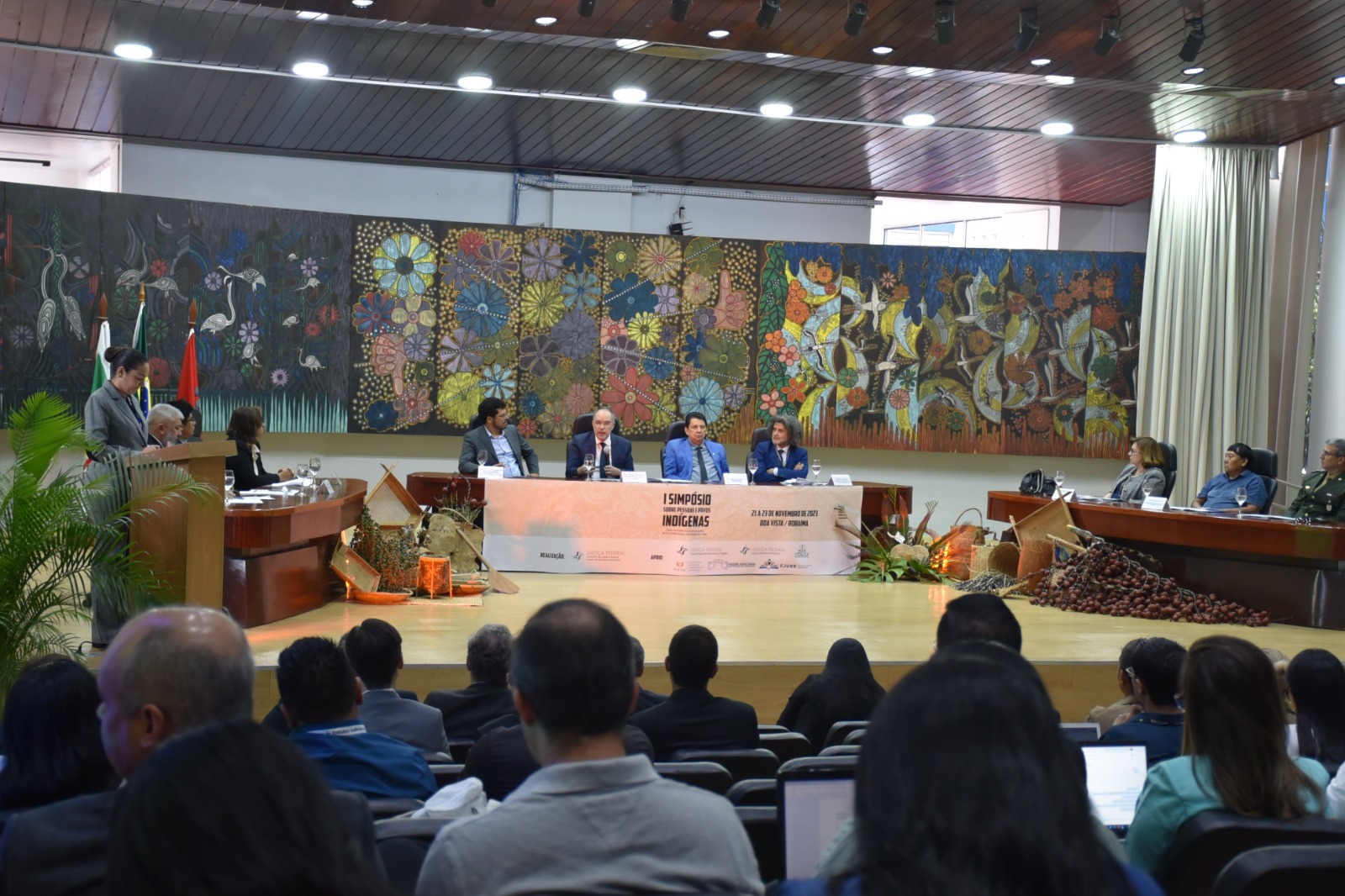 Foto colorida mostra uma plateia de costas para a câmera. A frente está o presidente do Poder Judiciário de Roraima, desembargador Jésus Nascimento, sentado ao lado do Corregedor Regional de Justiça Federal da 1ª Região, desembargador Néviton Guedes, que está com o microfone na mão, o juiz federal Erivaldo Santos. No fundo há um quadro com diversas flores coloridas. A frente há uma faixa escrito “ I Simpósio Sobre Pessoas e Povos Indígenas”, “21 a 23 de novembro de 2023, Boa Vista / Roraima”.