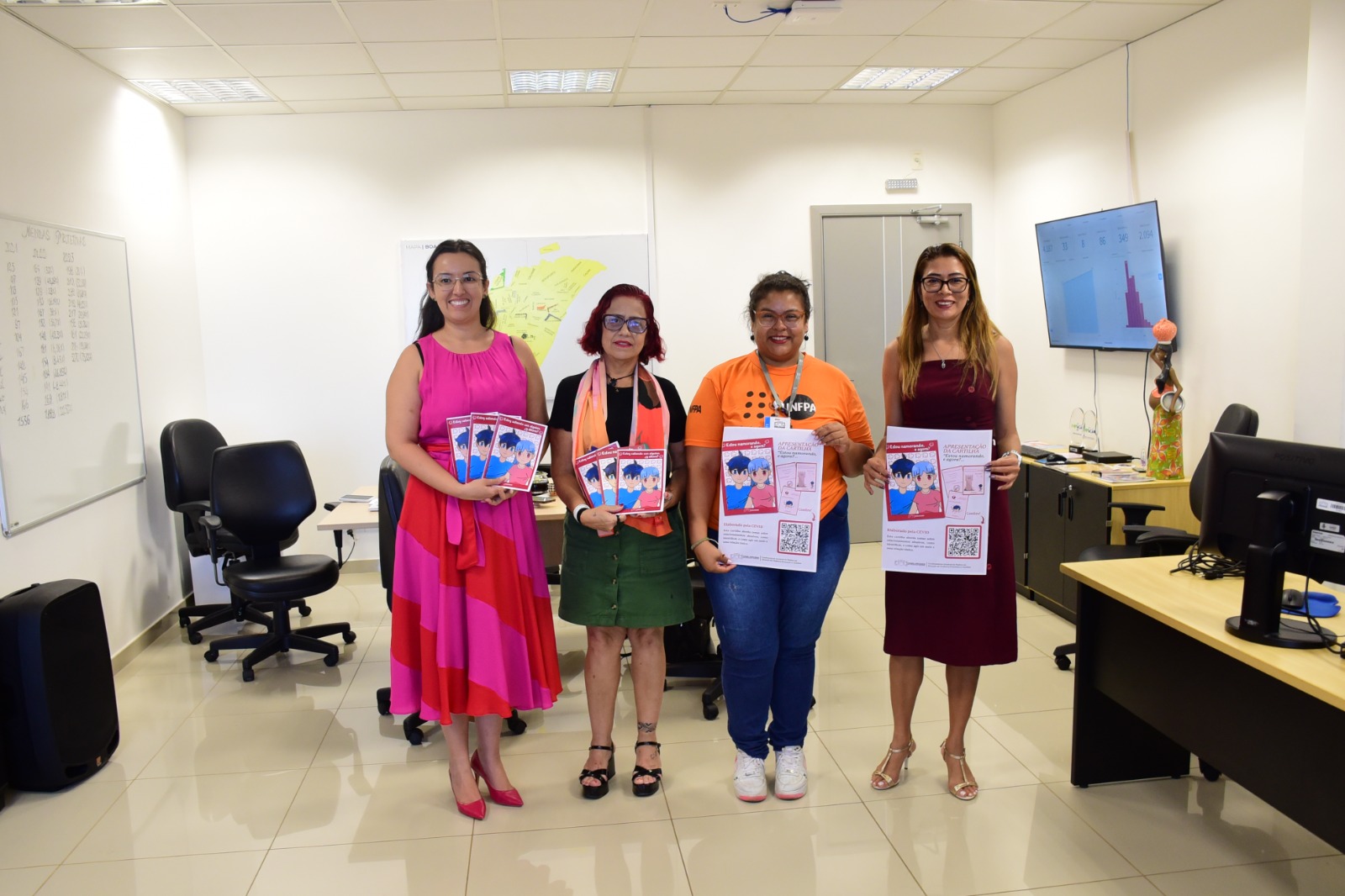 Imagem colorida mostra quatro mulheres segurando cartilhas informativas em uma sala ampla que tem algumas mesas e uma televisão ligada.