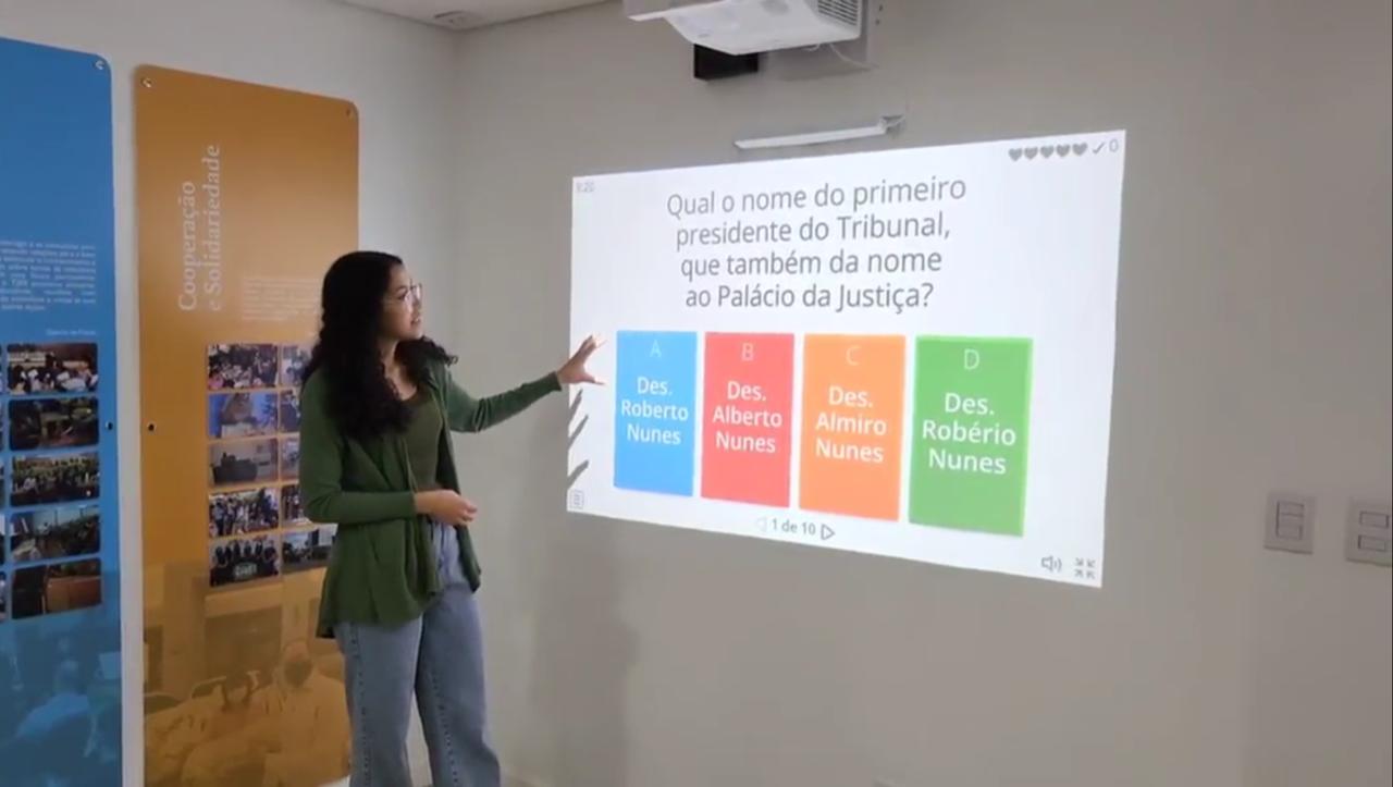 Imagem colorida mostra Imagem colorida mostra mulher apontando para slide exibido no projetor. Abaixo contém a frase “HISTÓRIA E INOVAÇÃO - Centro de Memória e Cultura do Poder Judiciário de Roraima instala projetor interativo a laser”.