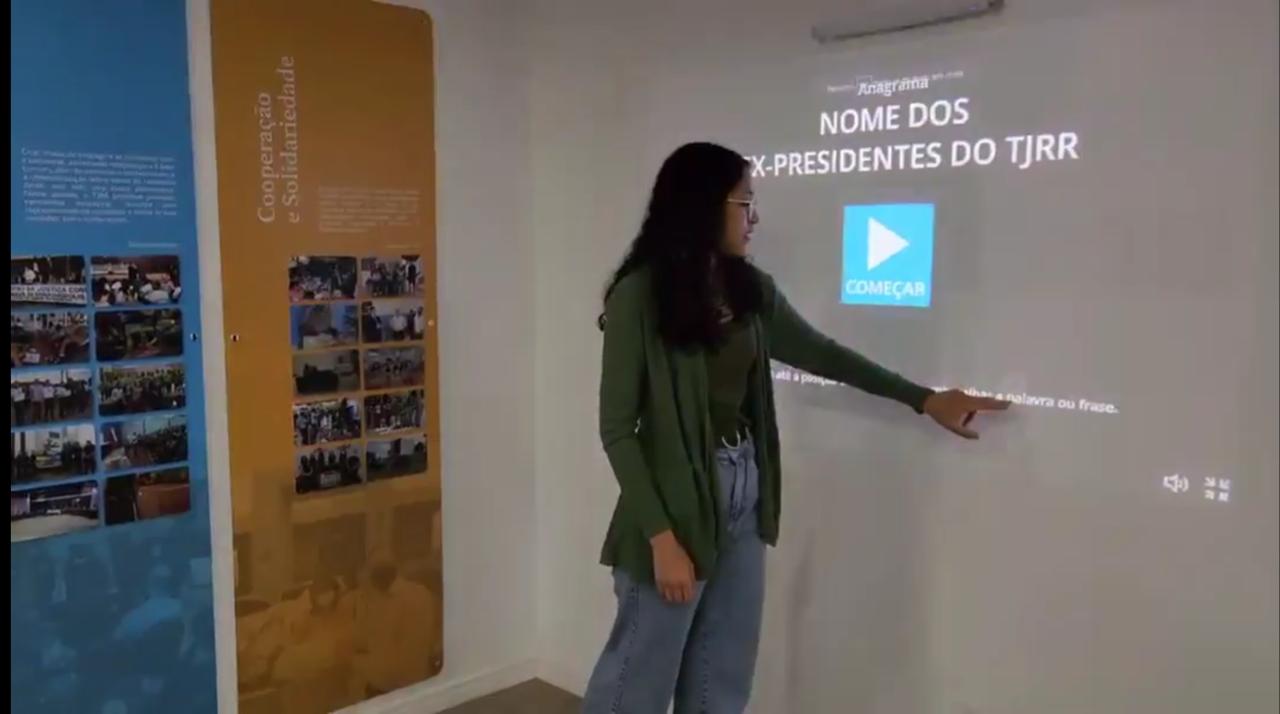 Imagem colorida mostra Imagem colorida mostra mulher apontando para slide exibido no projetor. Abaixo contém a frase “HISTÓRIA E INOVAÇÃO - Centro de Memória e Cultura do Poder Judiciário de Roraima instala projetor interativo a laser”.