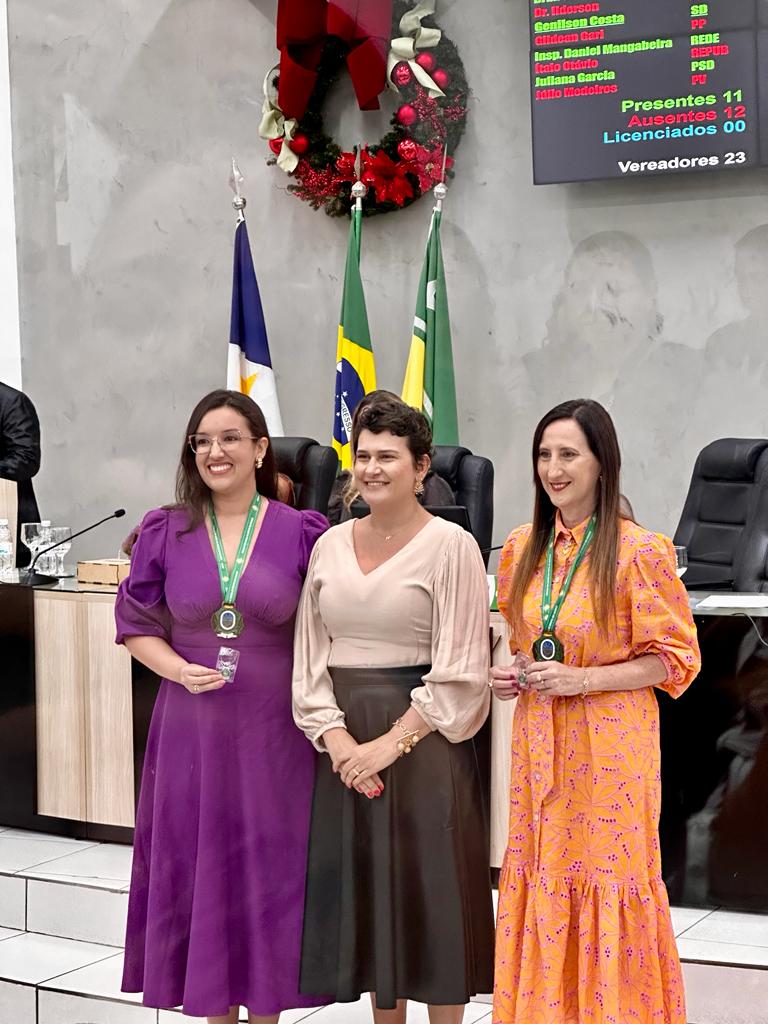 foto colorida da juíza na cerimônia com a medalha e segurando um broche ao lado de outra duas mulheres posando para a fotografia.