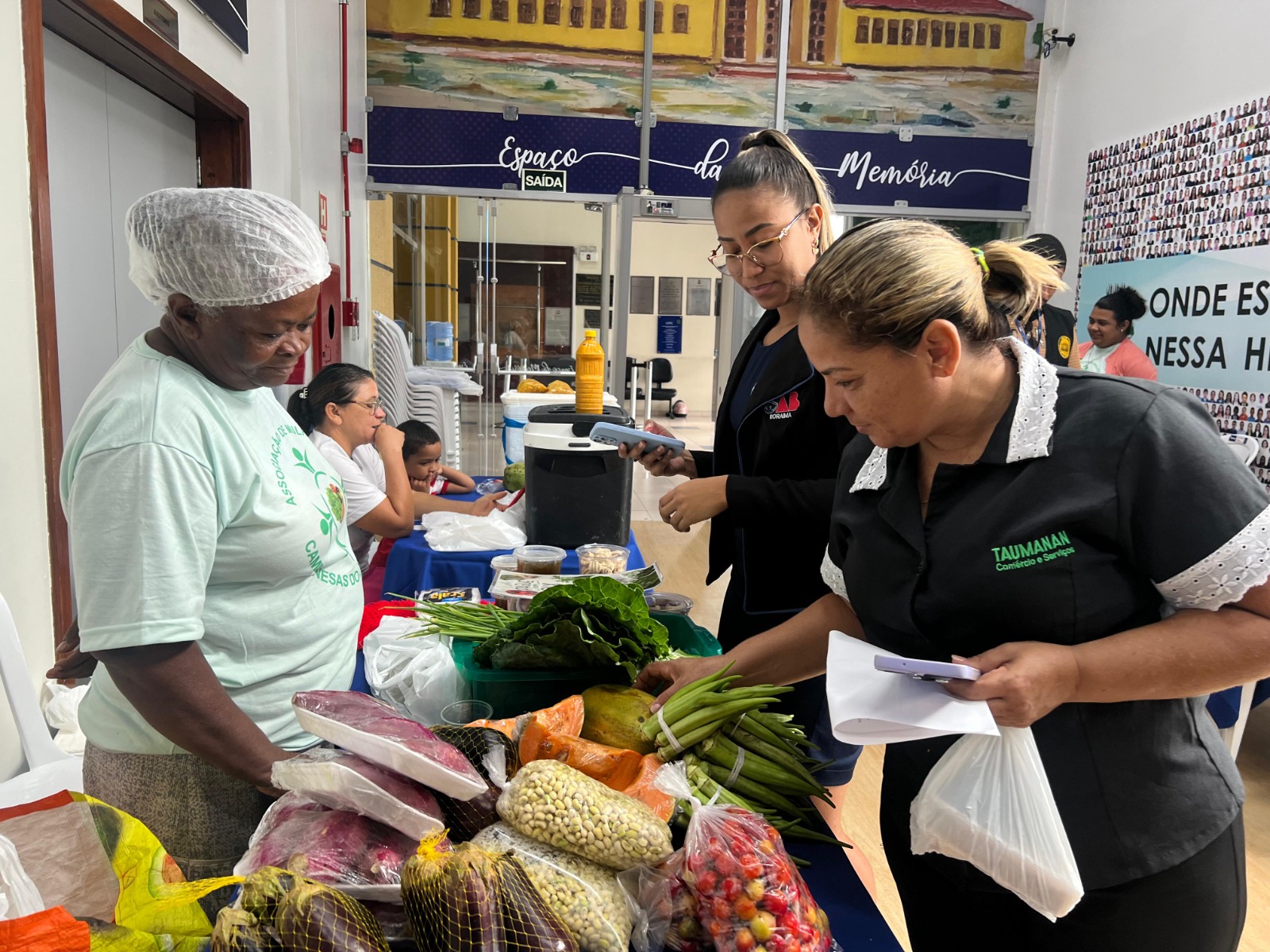 Imagem colorida mostra a Feira de orgânicos do TJRR, com a participação das Camponesas do Lavrado. Na imagem está ocorrendo uma venda de legumes, com seis pessoas na fotografia.
