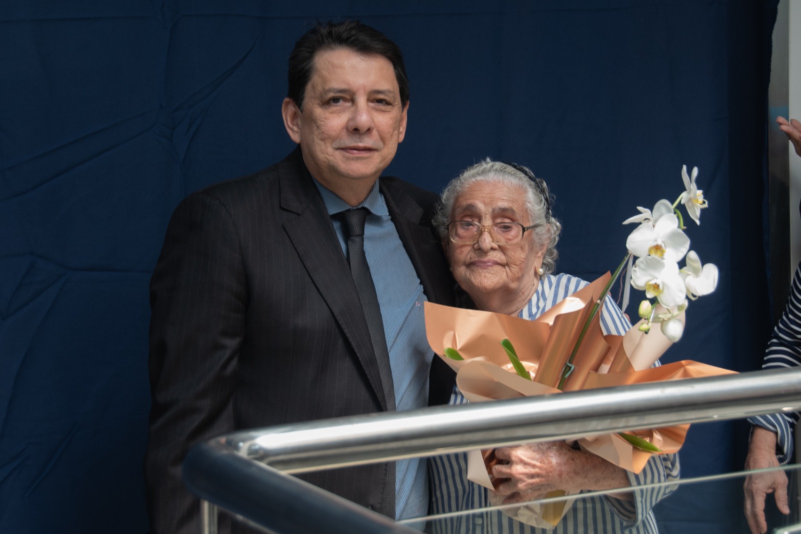 Imagem colorida mostra o Presidente do Tribunal de Justiça de Roraima, desembargador Jésus Nascimento, ao lado da a viúva do homenageado, Cristina Rocha, posando para foto com flores na mão.