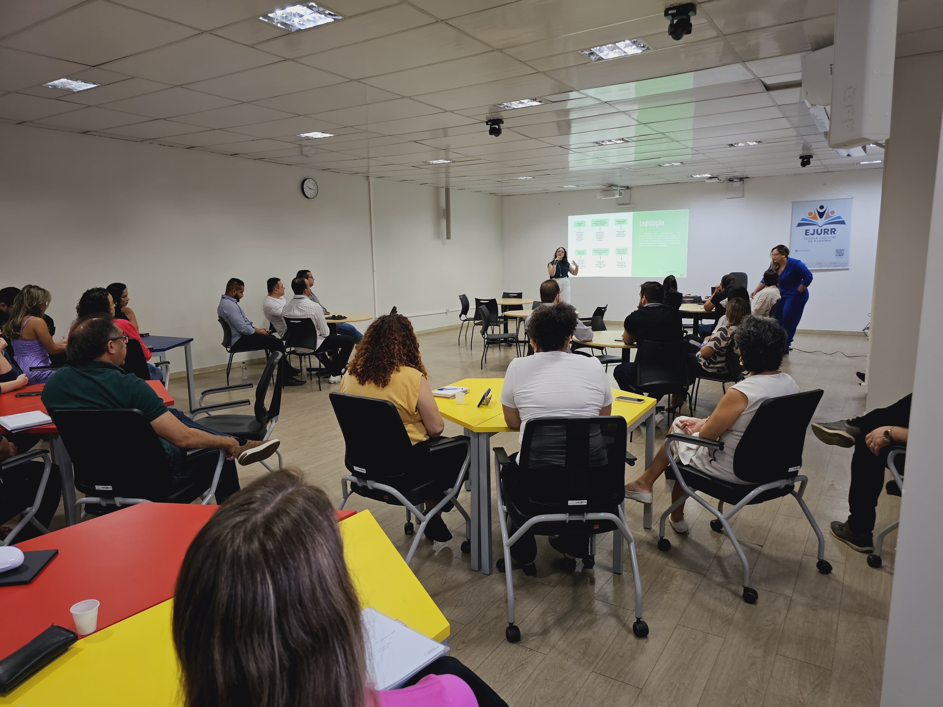 foto colorida de um espaço cheio de mesas compostas por equipes a frente há uma mulher falando no slide