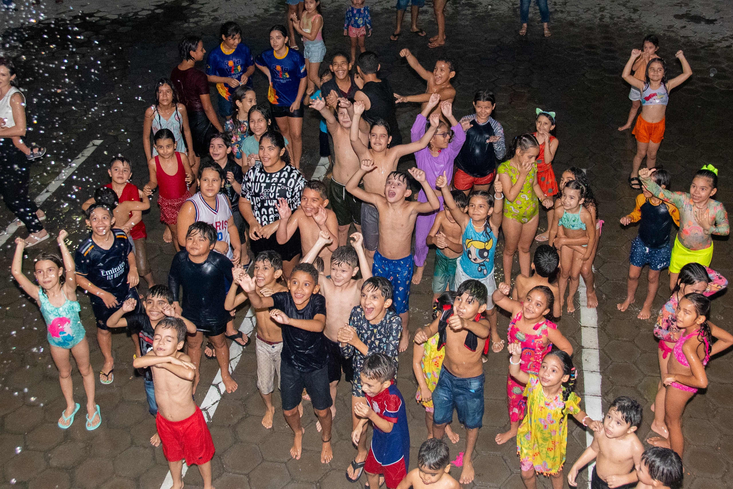 foto colorida em plano de cima que mostra um grupo de crianças felizes enquanto banham de mangueira do bombeiro