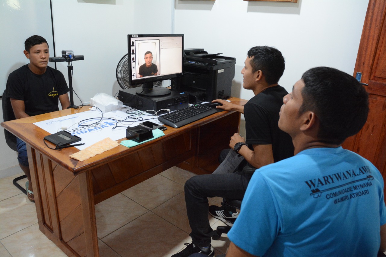 Imagem mostra dois indígenas em frente a um computador, atendendo um terceiro homem da comunidade. A foto dele aparece no computador para emissão de documento de identidade. 