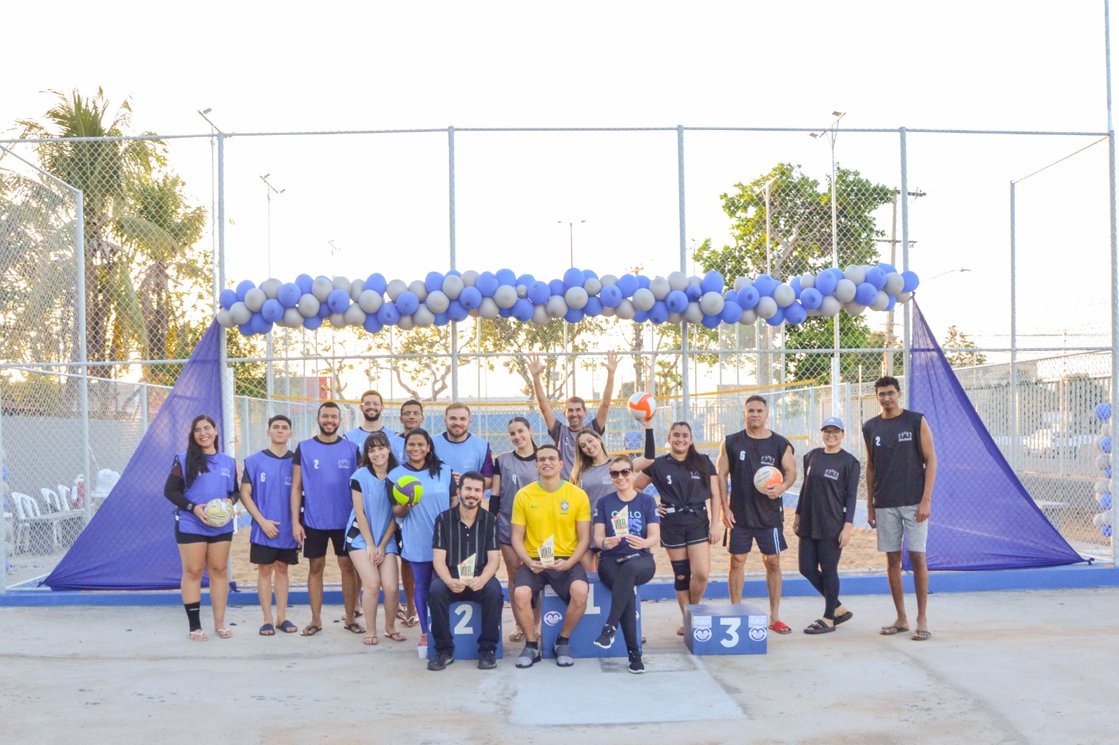 Imagem colorida mostrando pessoas posando para uma foto em frenta uma quadra de areia. As pessoas usando coletes esportivos e segurando bolas de volei