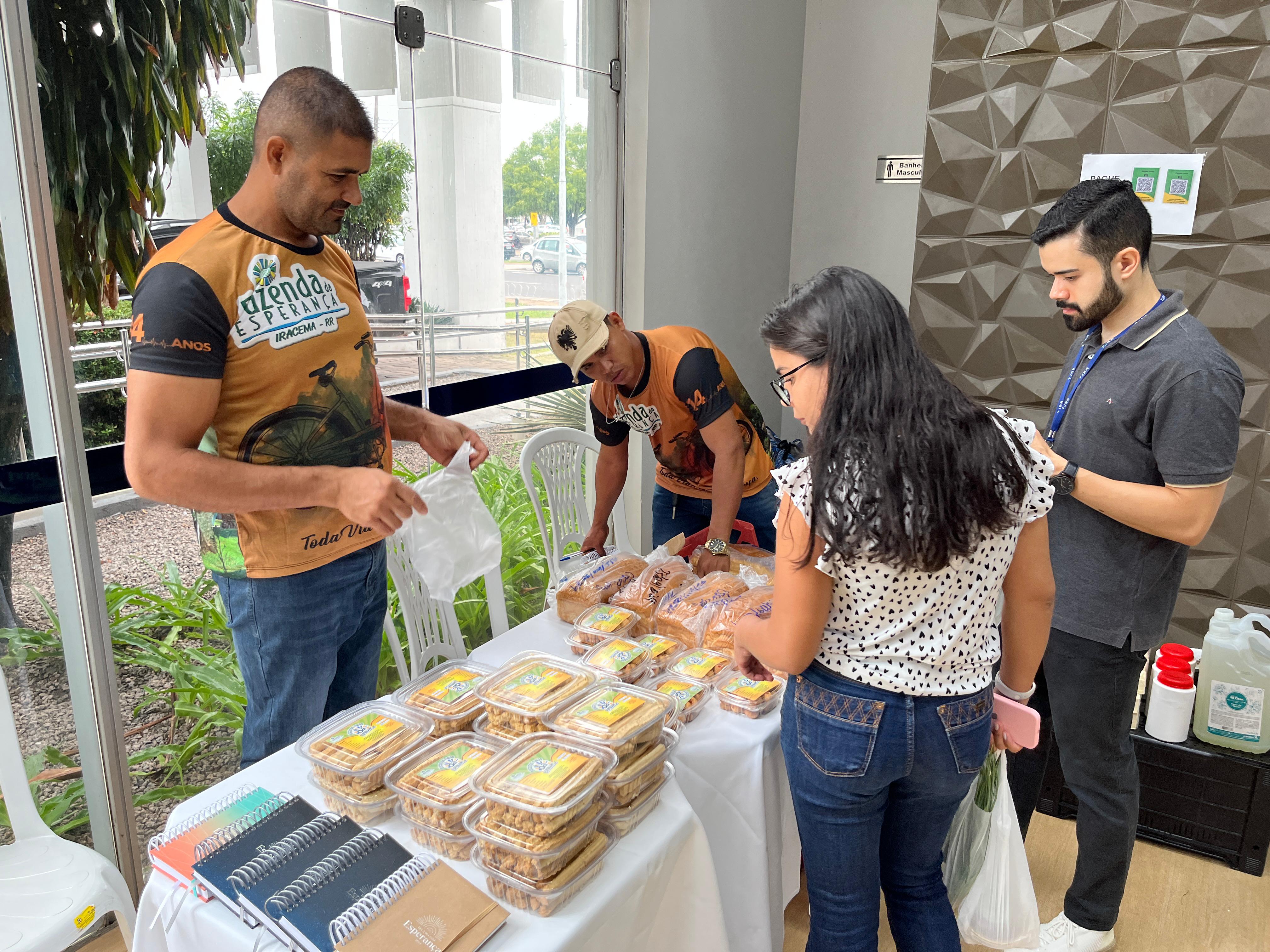 Foto colorida mostrando pessoas comprando produtos comercializados pela Fazenda da Esperança. Na mesa há pães e doces