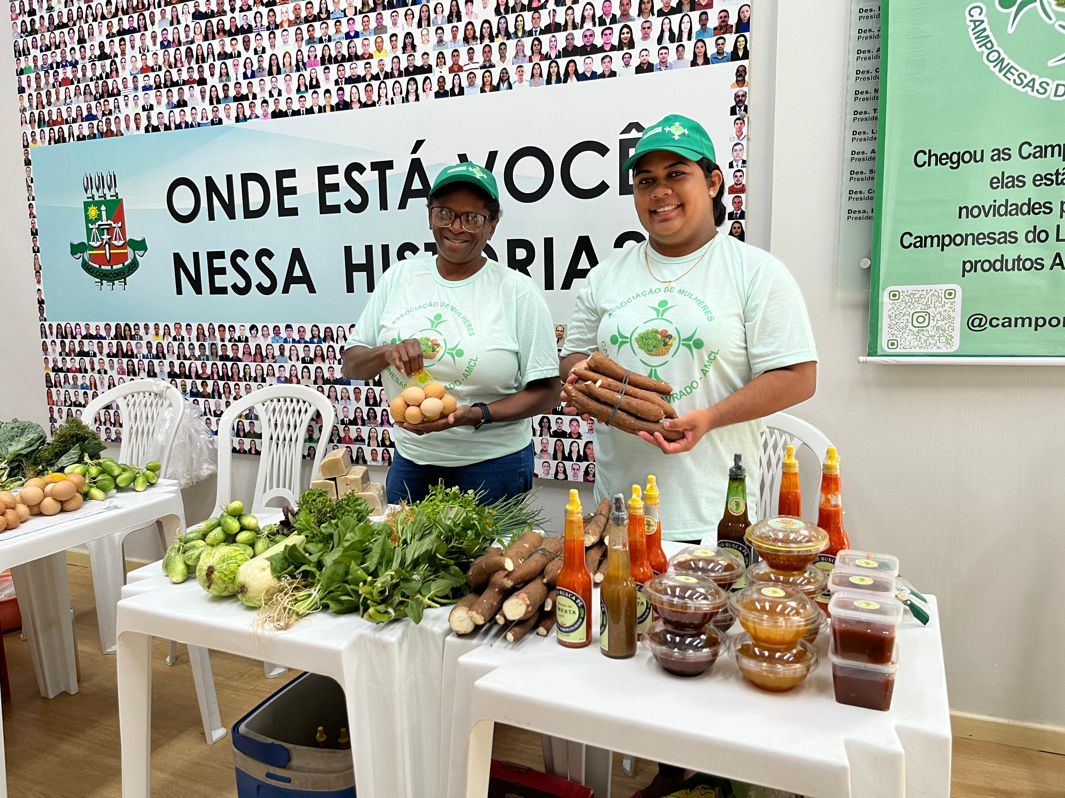 Imagem colorida mostrando trabalhadoras das Camponesas do Lavrado comercializando seus produtos. Na mesa, a frente delas, verduras, doces e potes de pimentas