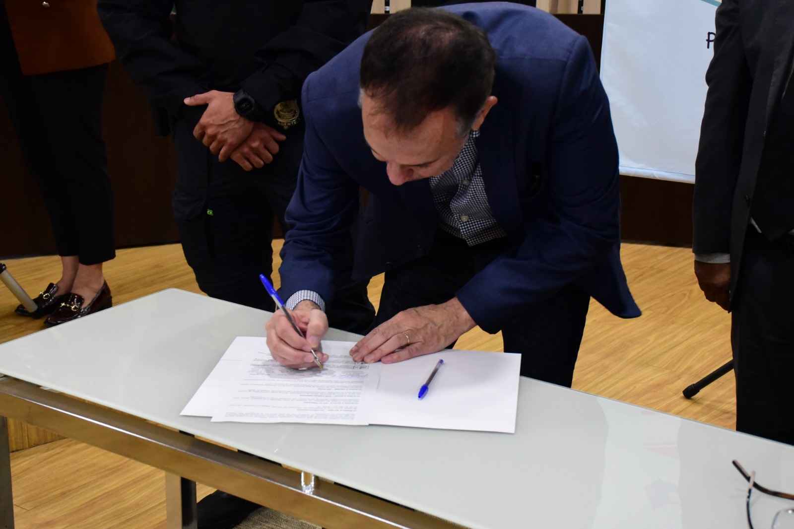 foto colorida dando enfoque no desembargador, supervisor do GMF, Almiro Padilha, assinando o acordo de cooperação entre o TJRR, Receita Federal e Sejuc que está em cima de uma mesa de vidro branco com um caneta bic azul.