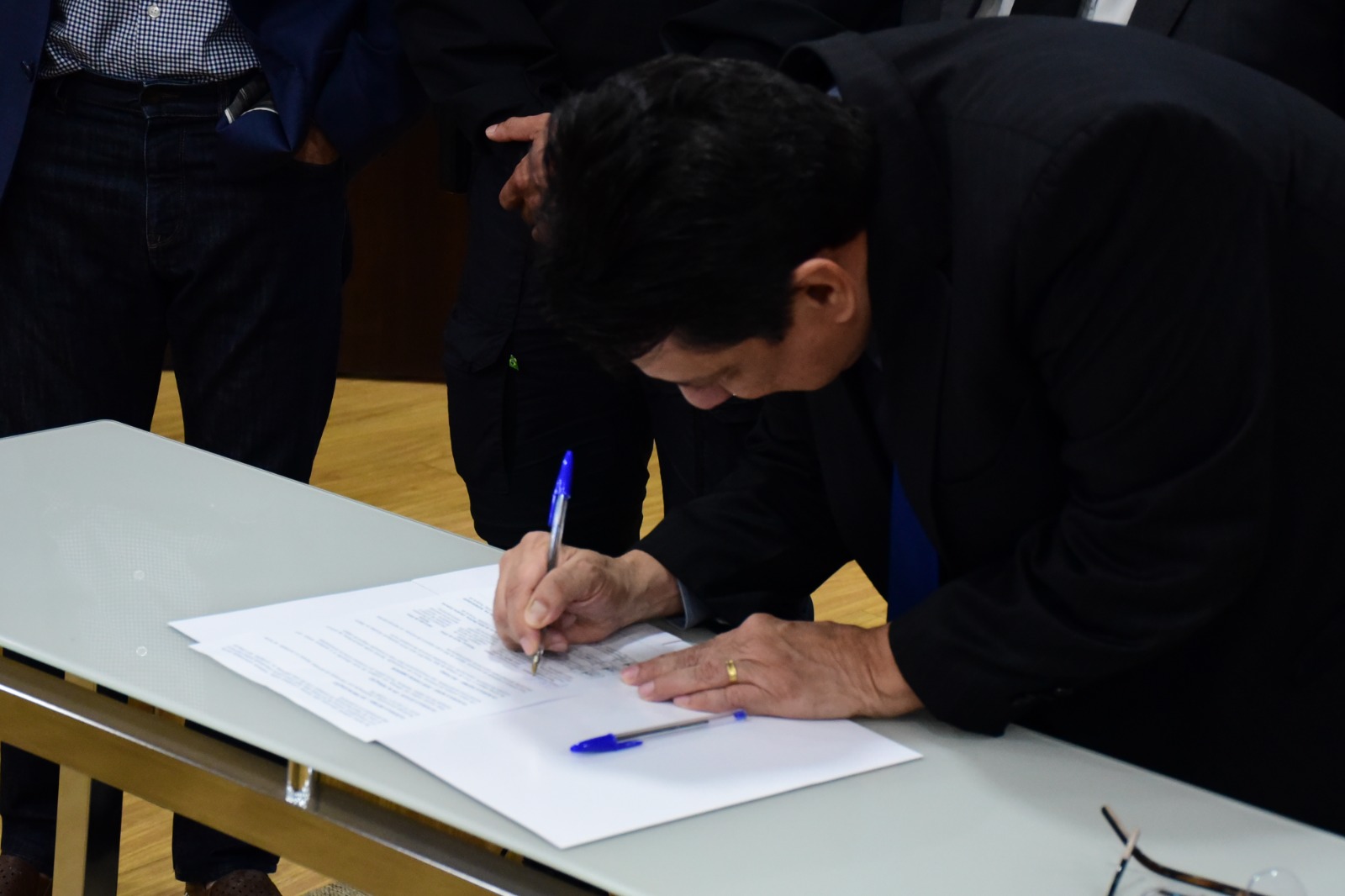 foto colorida dando enfoque no Presidente do TJRR, desembargador Jésus Nascimento, assinando o acordo de cooperação entre o TJRR, Receita Federal e Sejuc que está em cima de uma mesa de vidro branco com uma caneta bic azul.