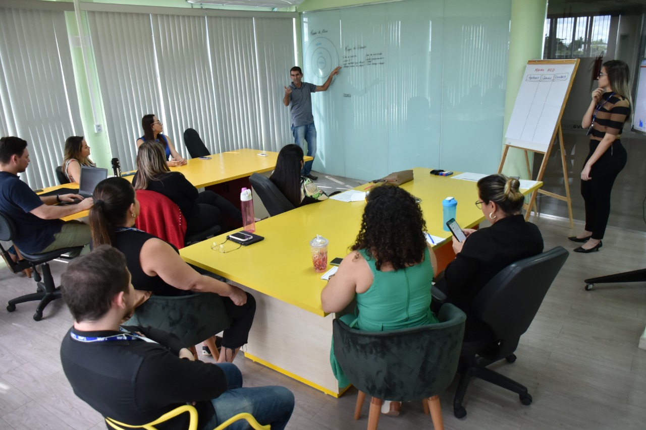 Imagem mostra uma reunião do Laboratório de Inovação. Os participantes estão em cadeiras pretas em torno de mesas amarelas e o instrutor está a frente deles fazendo anotações em um quadro branco.