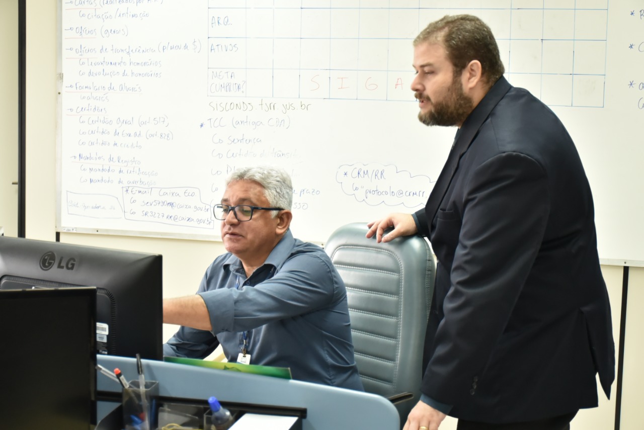 Imagem colorida contém o consultor Fábio Didier, responsável pela capacitação nos módulos de expansão do sistema de Gestão de qualidade da ISO 9001, auxiliando servidor do TJRR, que se encontra sentado de frente para o computador, com Fábio posicionado logo atrás. 