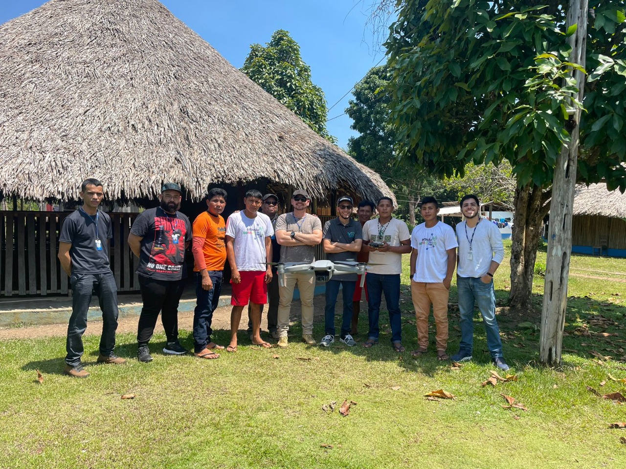  Imagem colorida mostra 11 homens e um drone pairando no ar, entre eles há  servidores do TJRR e indígenas da comunidade Waimiri-Atroari, os mesmos estão em pé posando para a foto em frente a um malocão.
