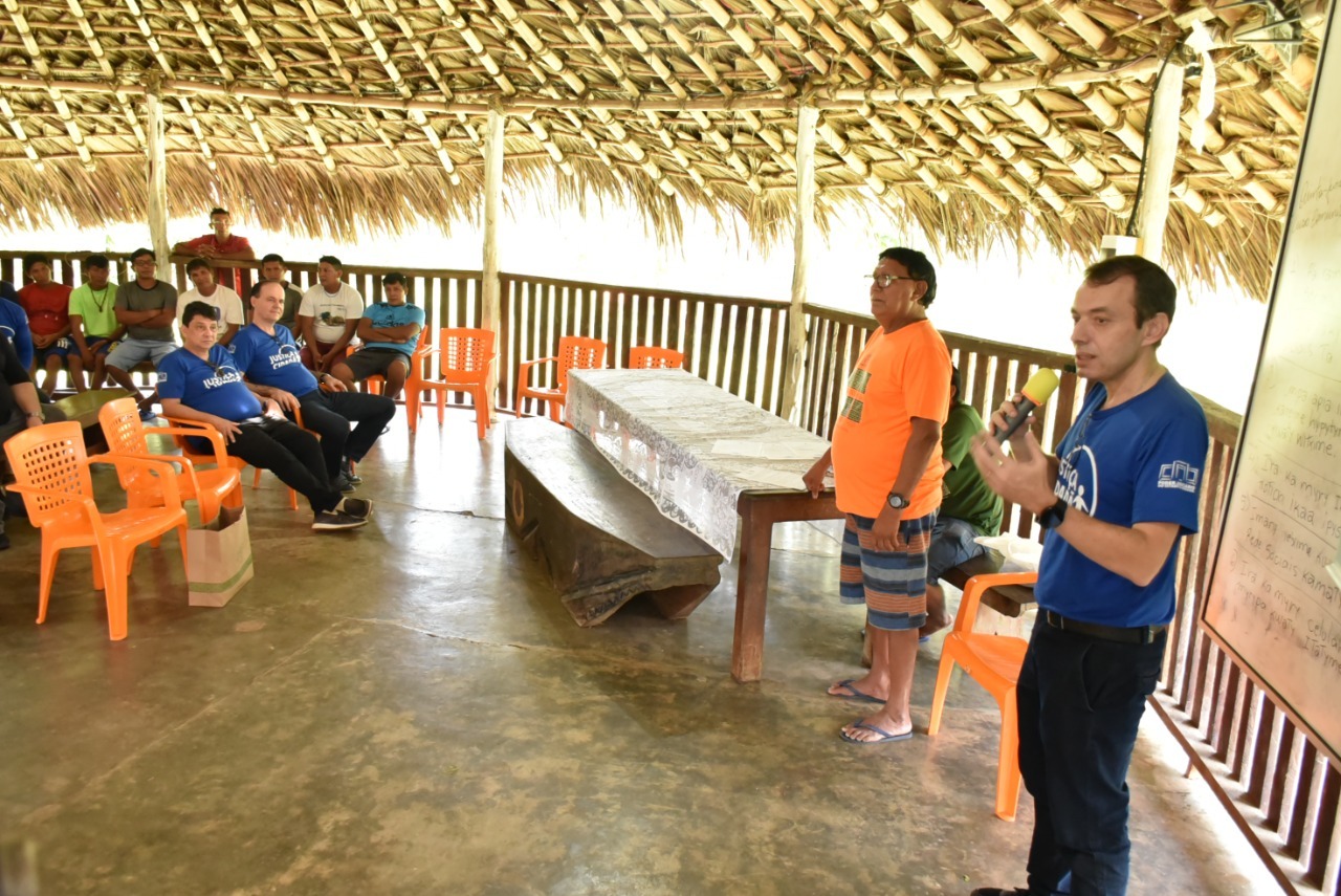 Imagem colorida em formato retângular mostra a área interna de um malocão. No canto esquerdo da foto está um pequeno público sentado prestigiando o evento. Na parte direita da foto está o desembargador Erick Linhares, discursando ao lado do líder indigena Mario Waimiri da comunidade Waimiri-Atroari.