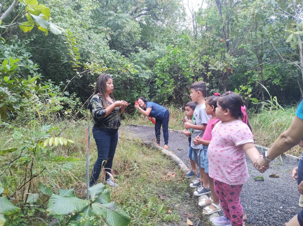 Imagem colorida ilustrativa mostra uma servidora conversando com as crianças, as mesmas estão de mãos dadas no limite da estrada de pedras, ao redor observa-se um fundo com diversos tipos de vegetação.