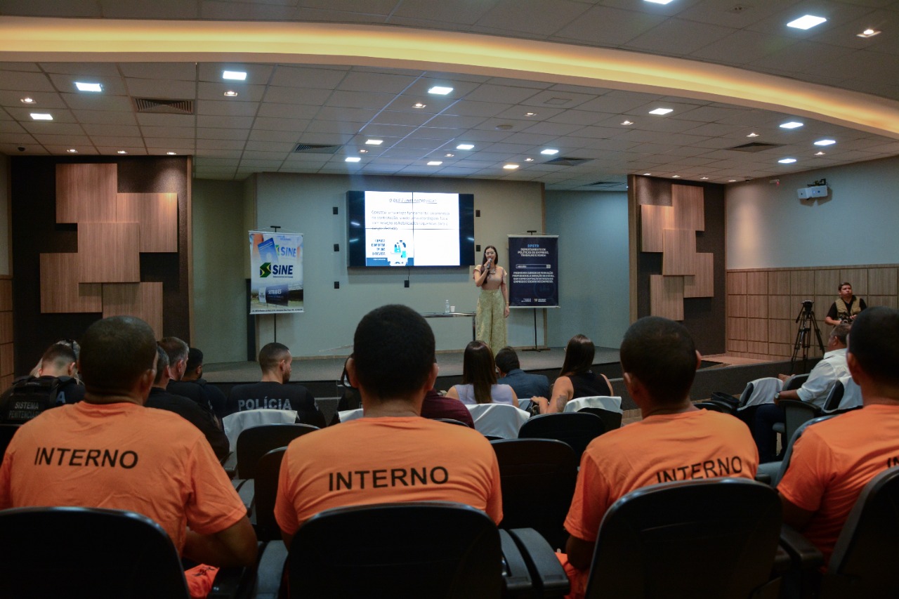 Imagem colorida mostra quatro internos do sistema prisional vestidos de laranja e um grupo de policiais e servidores sentados em um auditório, à frente está a palestrante em pé com um microfone na mão.  