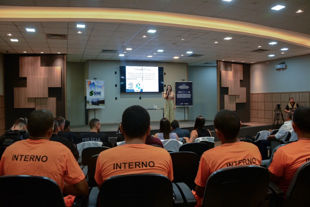 Imagem colorida mostra quatro internos do sistema prisional vestidos de laranja e um grupo de policiais e servidores sentados em um auditório, à frente está a palestrante em pé com um microfone na mão.