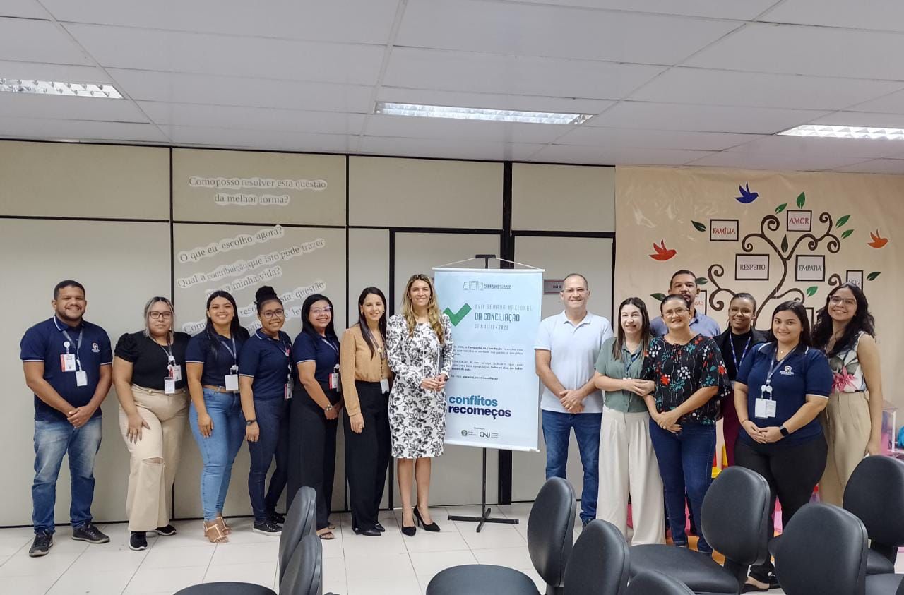 Imagem colorida mostra 11 mulheres e três homens posando em pé para uma foto, no centro da imagem há um banner com fundo branco com informações sobre a 17ª edição da semana estadual de conciliação, ao fundo há uma parede no tom bege com frases e desenhos coloridos simulando galhos de árvores com pássaros coloridos ao redor.  