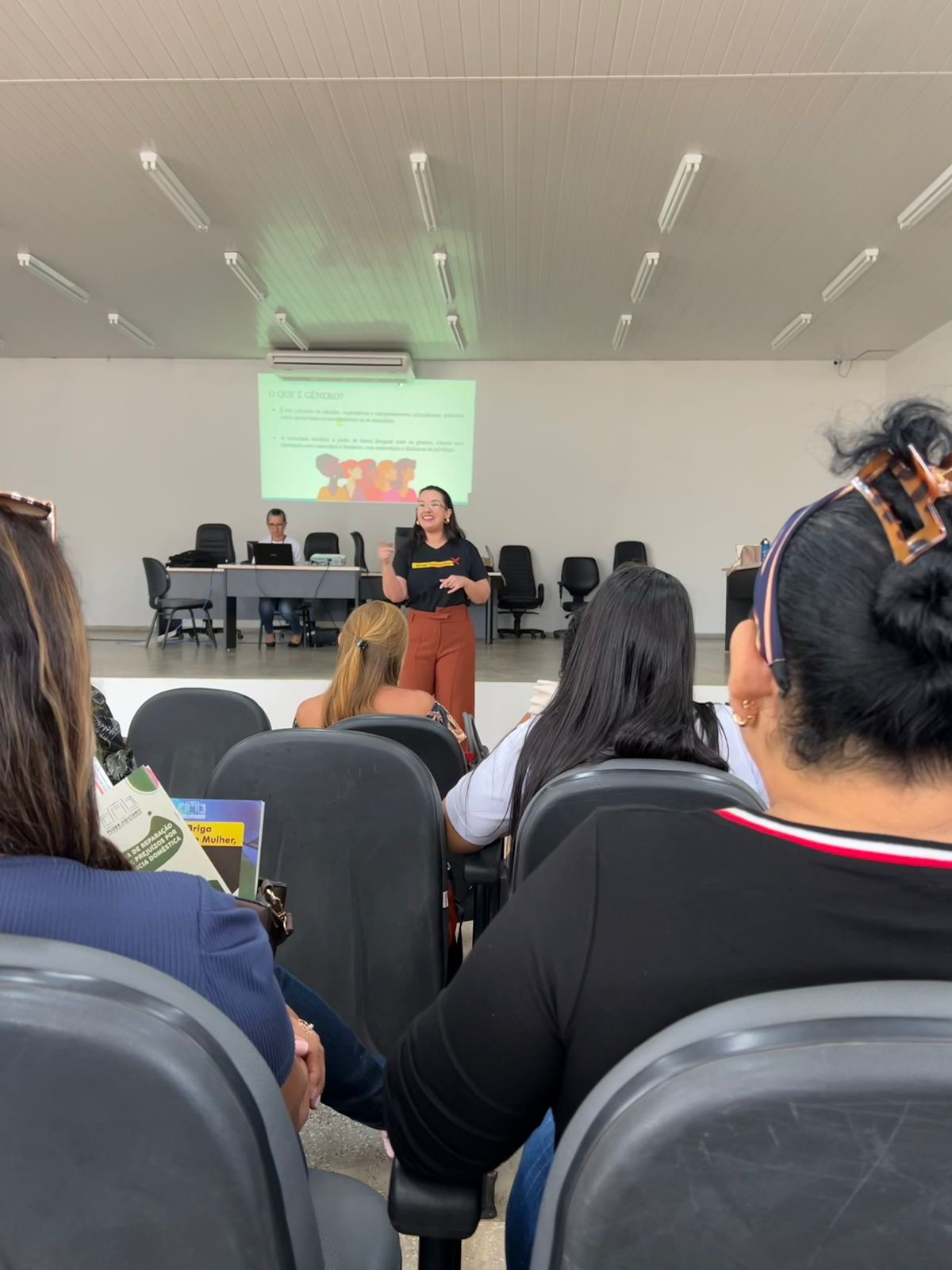 Imagem colorida em formato retangular mostra a coordenadora estadual de Violência Doméstica do Tribunal de Justiça de Roraima, juíza Suelen Alves palestrando para um grupo de mulheres em um auditório.