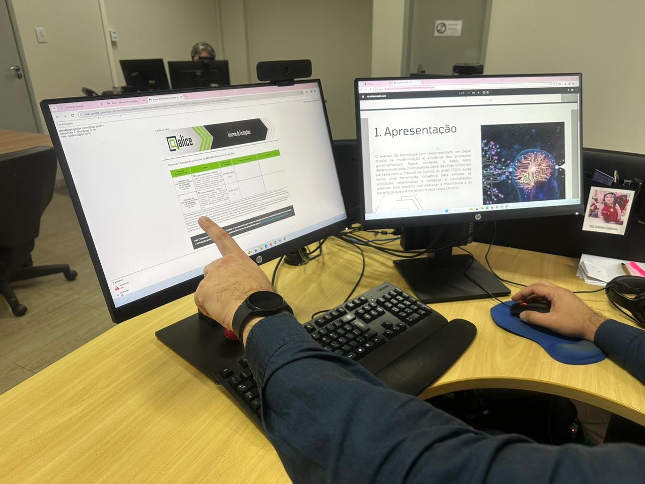 Foto colorida mostra dois monitores em cima de uma mesa, com uma mão apontando para o monitor da esquerda, que mostra o robô "Alice".