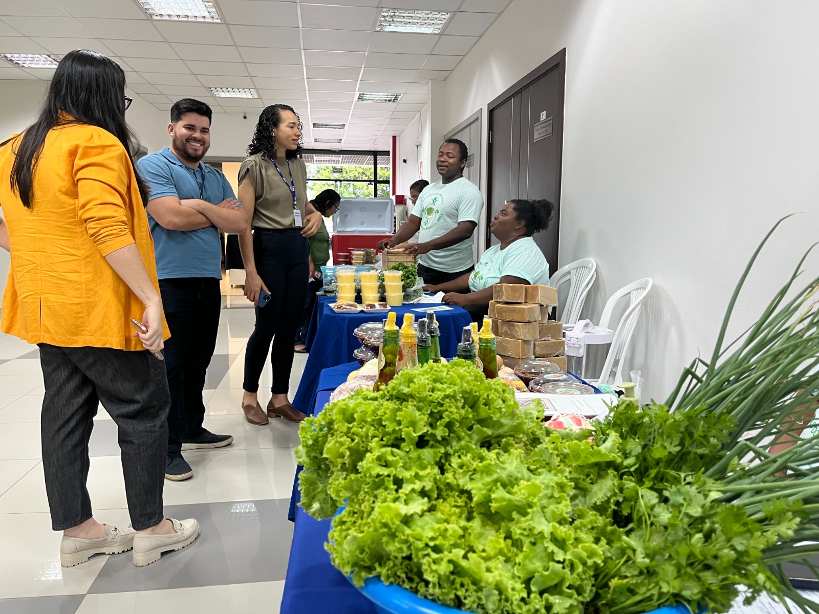 Foto colorida mostra à esquerda, 3 servidores olhando os produtos, que se encontram na mesa à direita da imagem.. Mais no fundo encontra-se 2 representantes da Associação Camponesas do Lavrado.