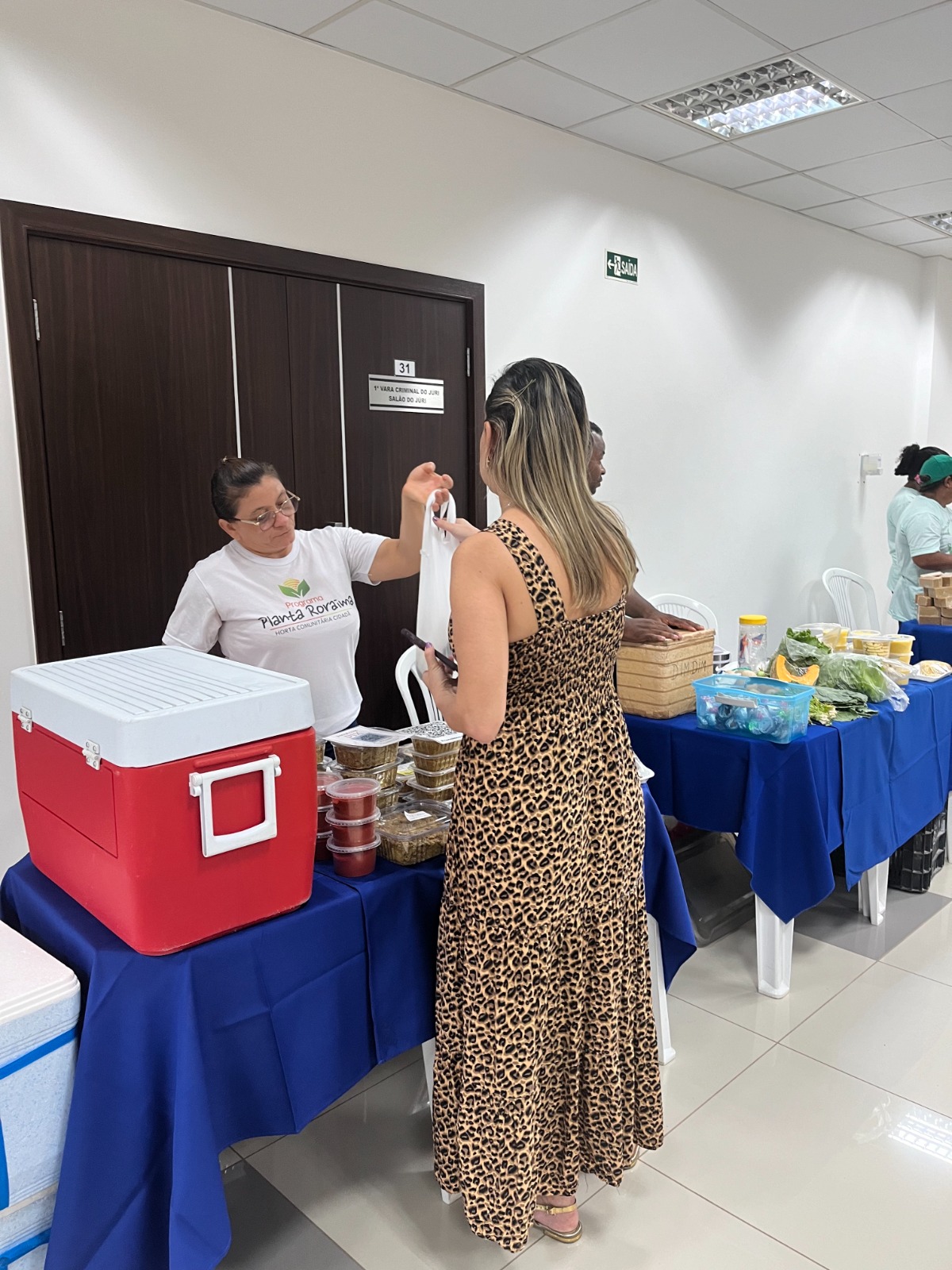Foto colorida mostra uma das empreendedoras presentes vendendo seu produto para uma servidora presente, que está recebendo uma sacola com esse produto.