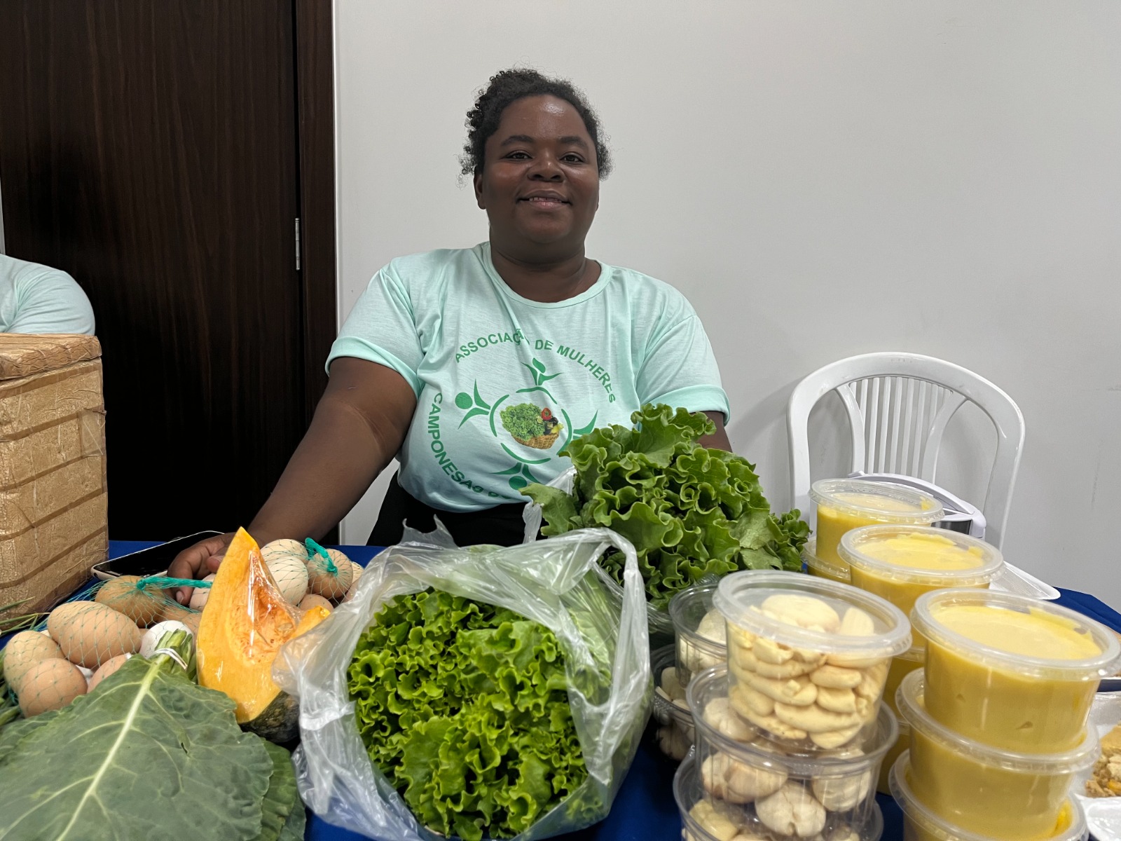 Foto colorida mostra uma das representados das Camponesas do Lavrado, Mirian Castro, que está posando para foto atrás da mesa com produtos orgânicos em cima.