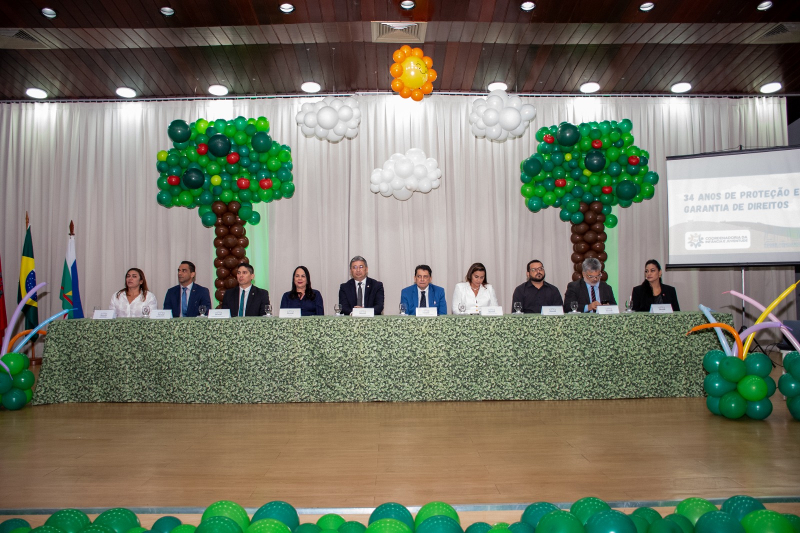imagem colorida mostra o presidente do TJRR, desembargador Jésus Nascimento, sentado na mesa de autoridades, durante evento no Fórum Cível, Advogado Sobral Pinto. Ao lado dele está autoridades do Estado de Roraima. Todos vestidos de roupas sociais. Atras deles encontram-se várias bexigas de balão de ar montados em formato de árvores, nuvens e um sol, todos de bexiga de ar.
